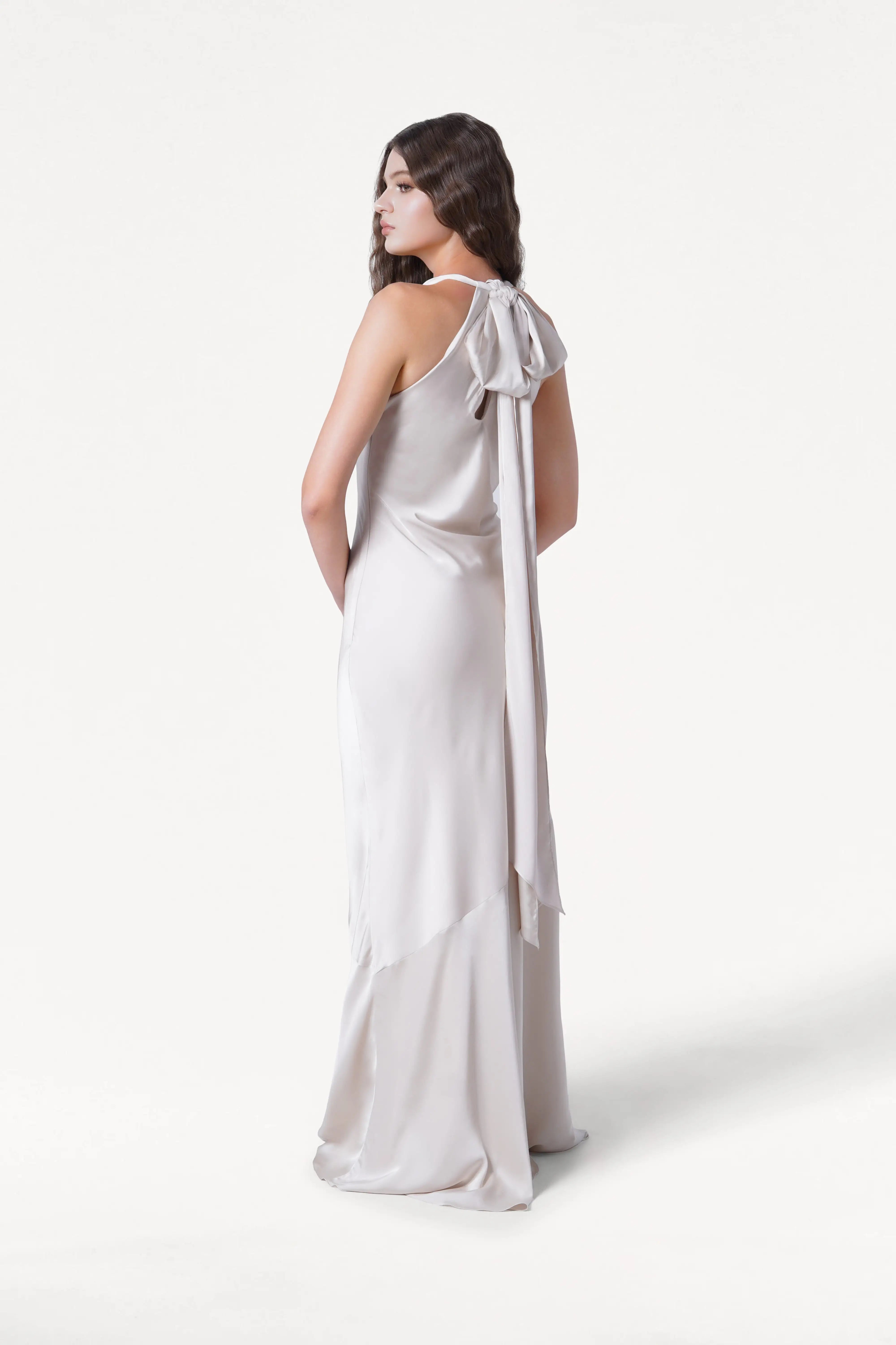 A woman models the Mira Bridesmaid Dress in Oyster Satin by TH&TH, featuring a long, sleeveless design with a large back bow and sash. She stands against a white background, looking over her shoulder with her hands in her pockets.