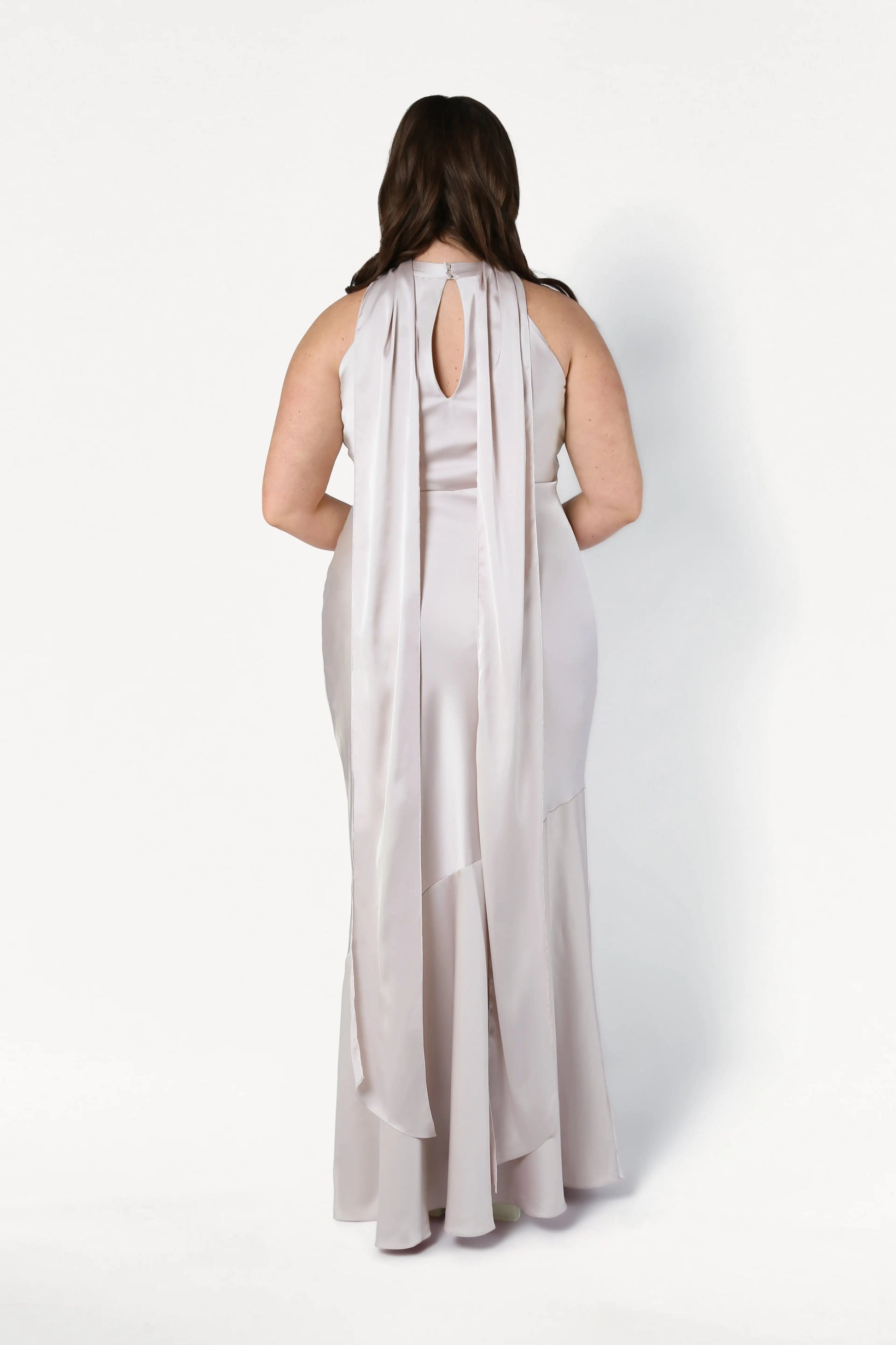 A woman in the TH&TH Mira Bridesmaid Dress in Oyster Satin stands with her back to the camera, showcasing its long, sleeveless design with a keyhole upper back and two flowing panels draping down.
