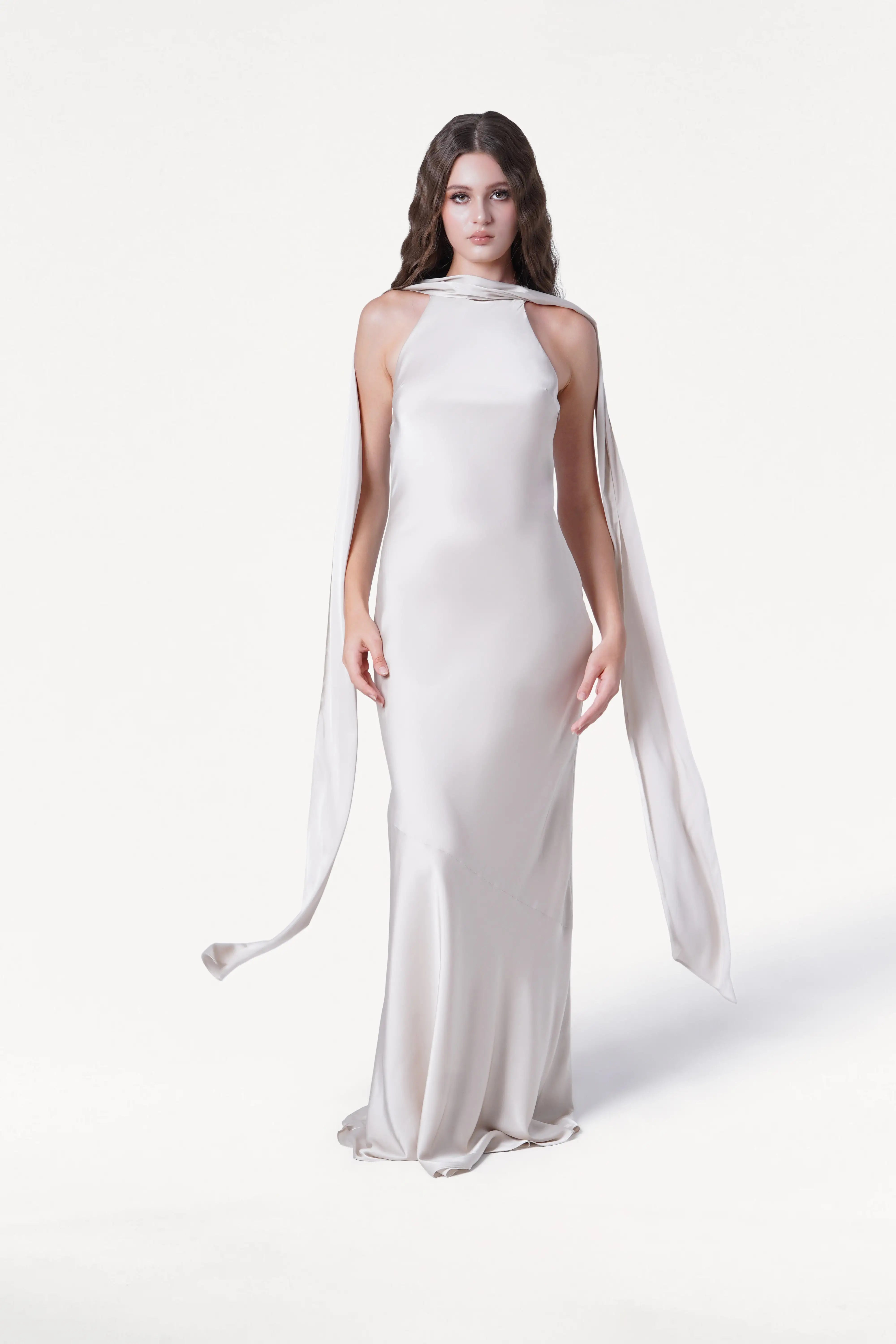 A woman with long wavy hair wears the Mira Bridesmaid Dress in Oyster Satin by TH&TH, featuring a halter neck and scarf-like ties. She stands against a plain white background.
