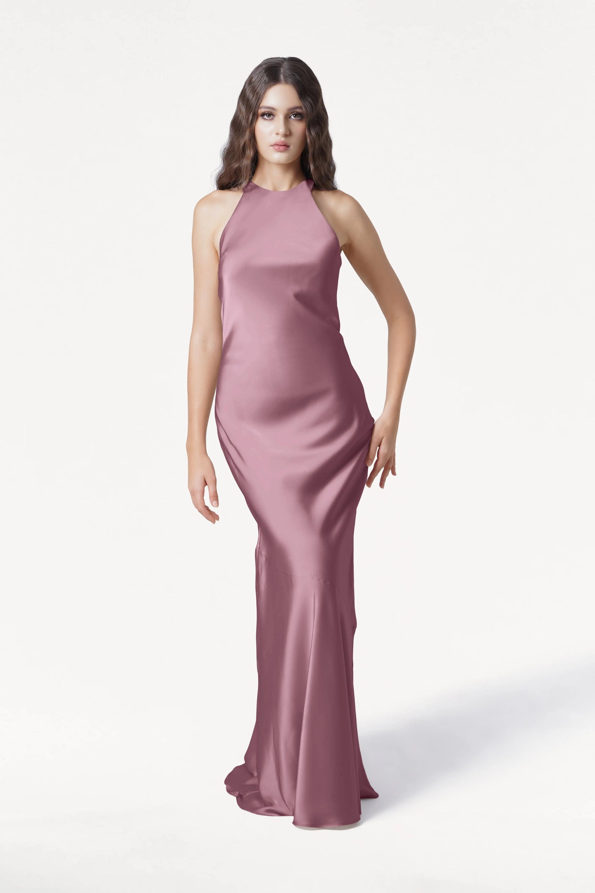 A woman with wavy brown hair models the Mira Bridesmaid Dress in Rose Pink Satin by TH&TH, a sleeveless, floor-length gown, standing against a plain white background.