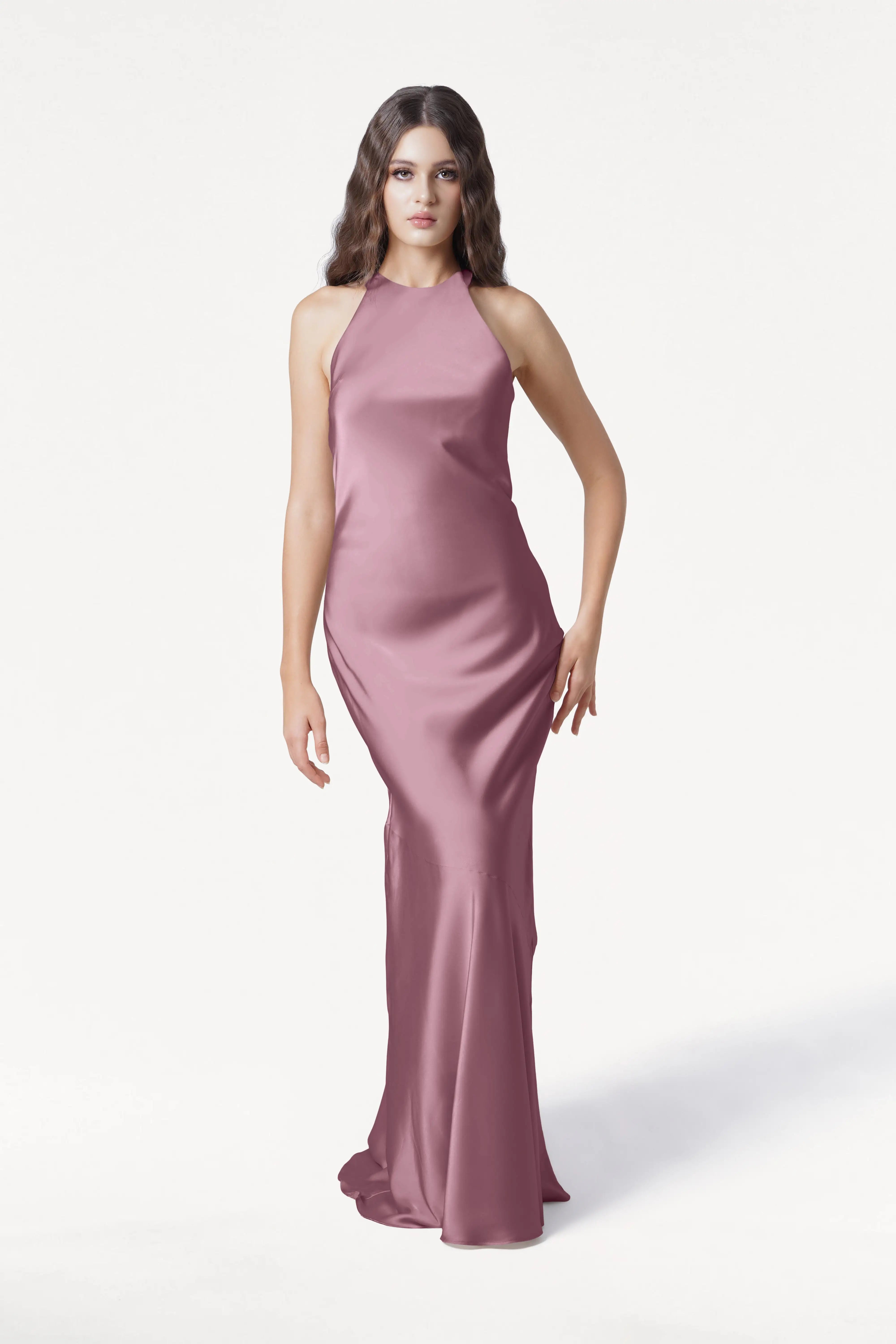 A woman with wavy brown hair models the Mira Bridesmaid Dress in Rose Pink Satin by TH&TH, a sleeveless, floor-length gown, standing against a plain white background.