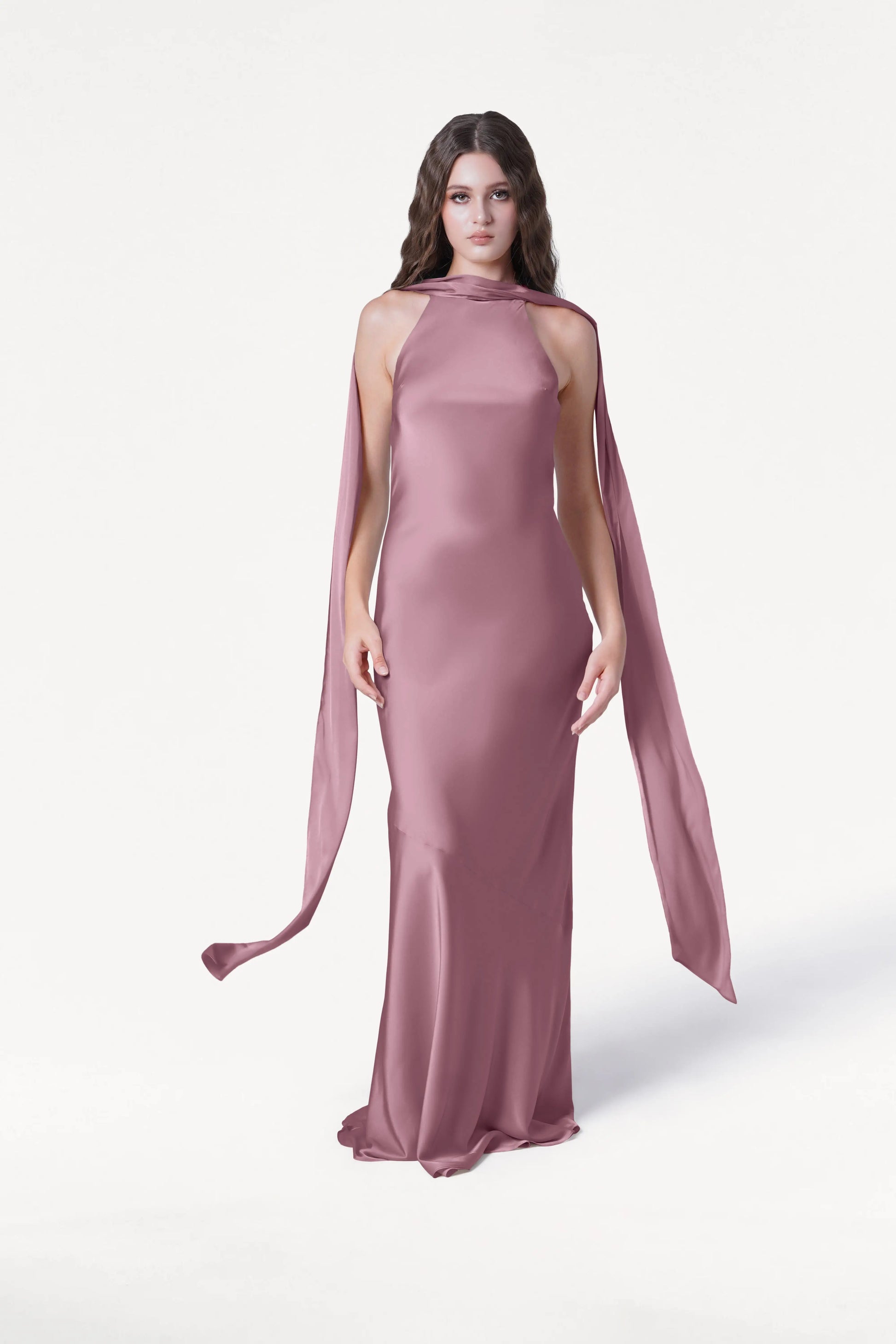 A woman with long brown hair models the TH&TH Mira Bridesmaid Dress in Rose Pink Satin, featuring a form-fitting bias-cut silhouette, high neckline, and flowing scarf detail, standing against a plain white background.