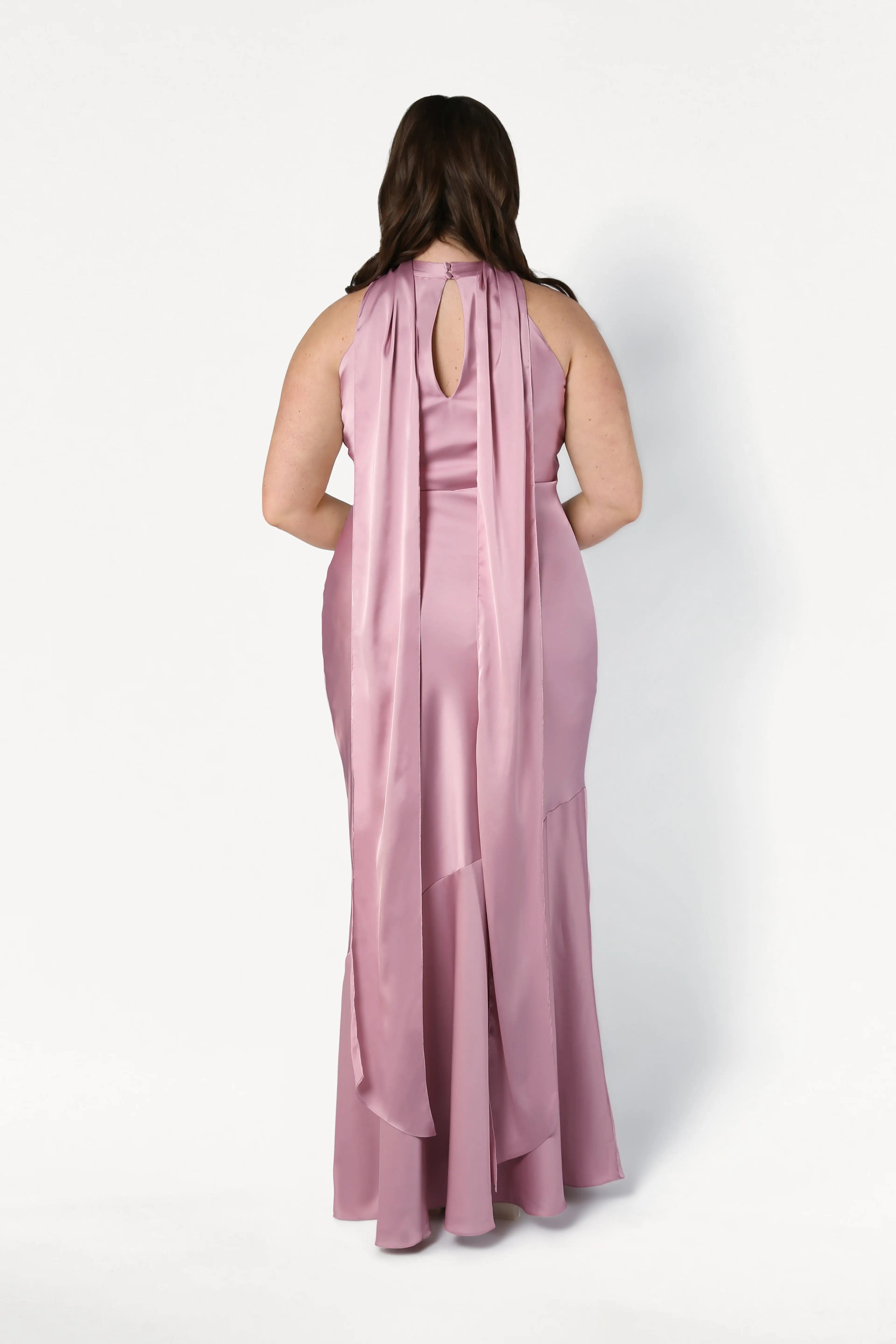 A woman with long brown hair models the TH&TH Mira Bridesmaid Dress in Rose Pink Satin, featuring a bias-cut silhouette, draped back, and flowing train, standing against a plain white background.