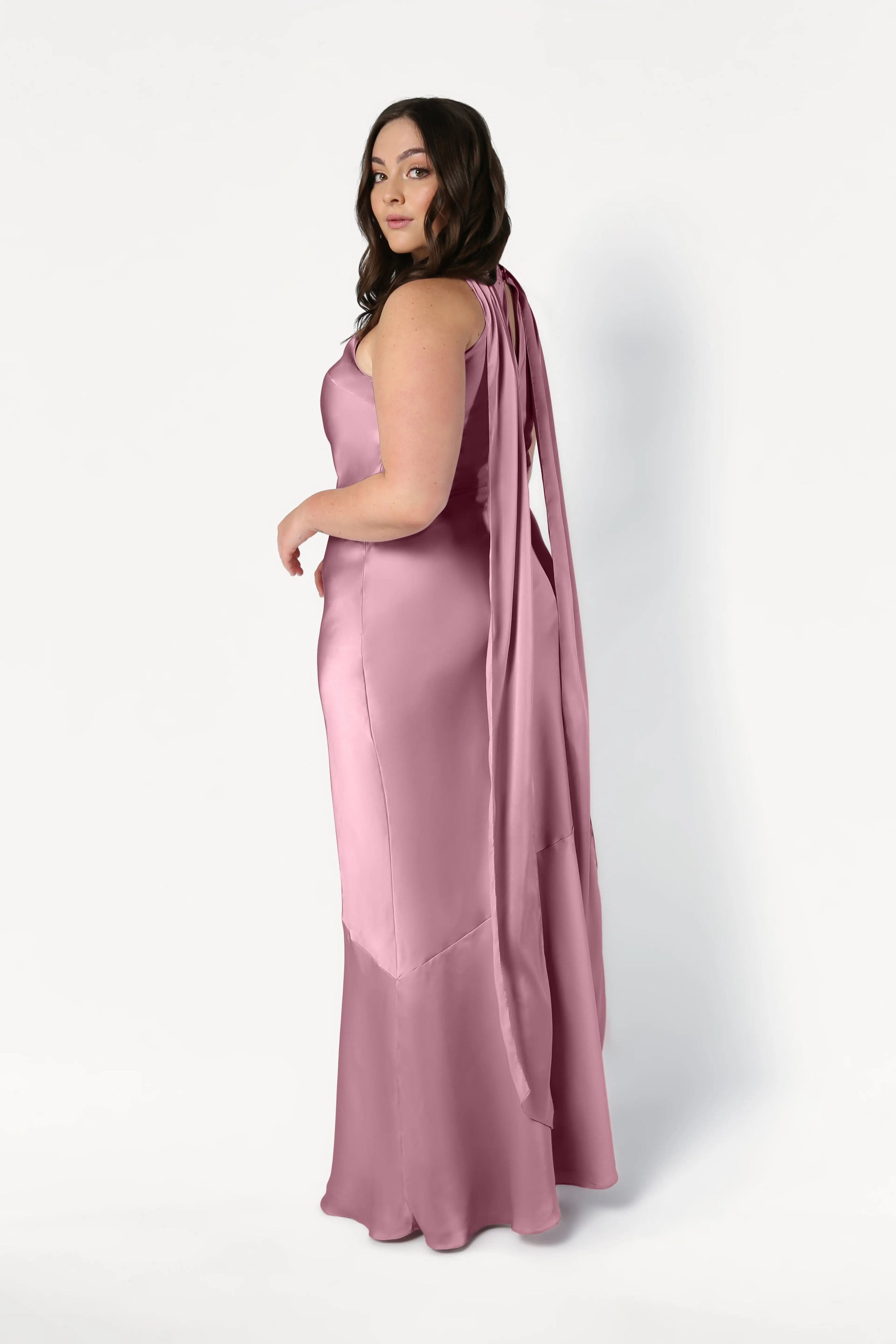 A woman with long brown hair poses in the Mira Bridesmaid Dress by TH&TH, a sleeveless, floor-length rose pink satin gown with a draped back and bias-cut silhouette, looking over her shoulder against a plain white background.