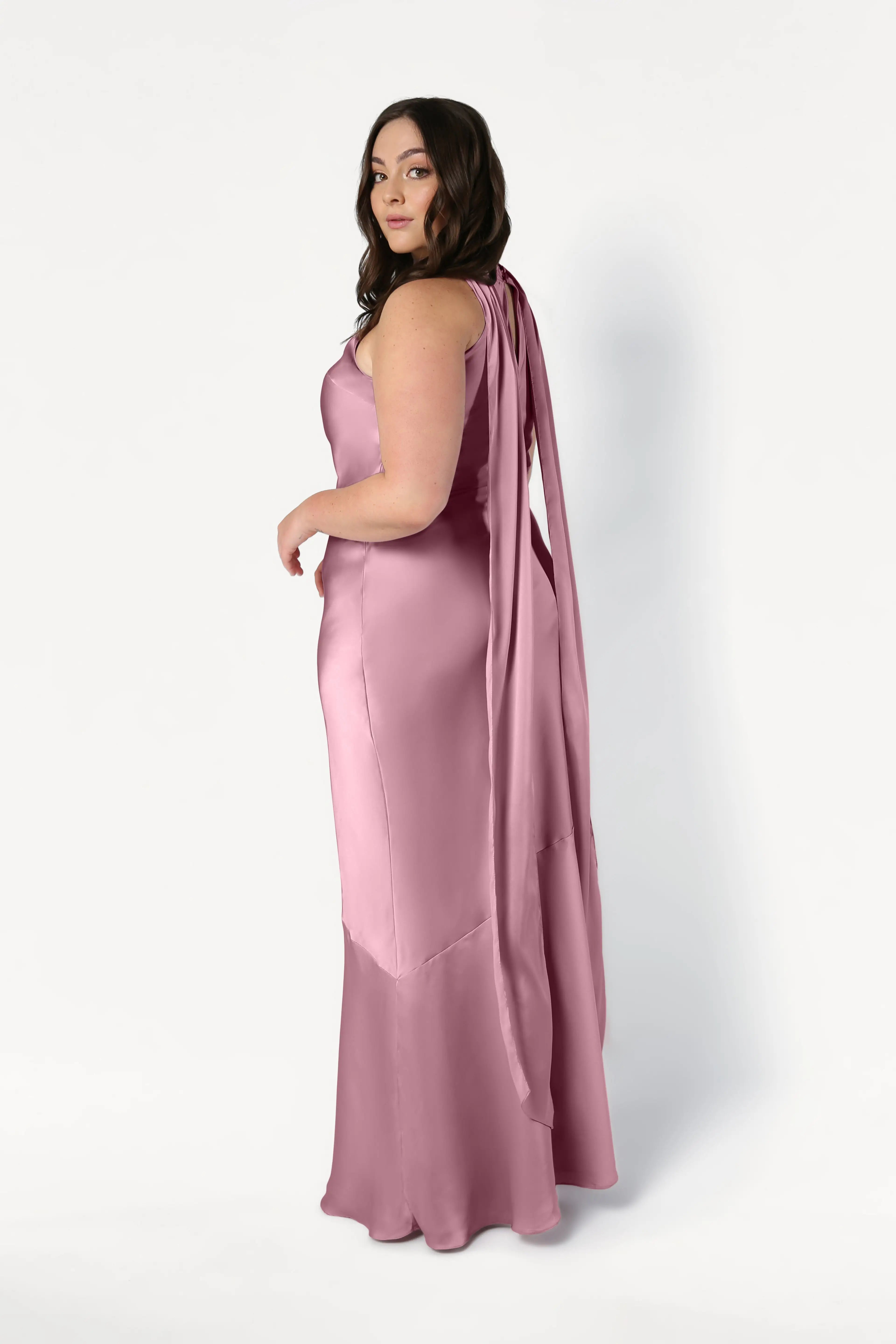 A woman with long brown hair poses in the Mira Bridesmaid Dress by TH&TH, a sleeveless, floor-length rose pink satin gown with a draped back and bias-cut silhouette, looking over her shoulder against a plain white background.