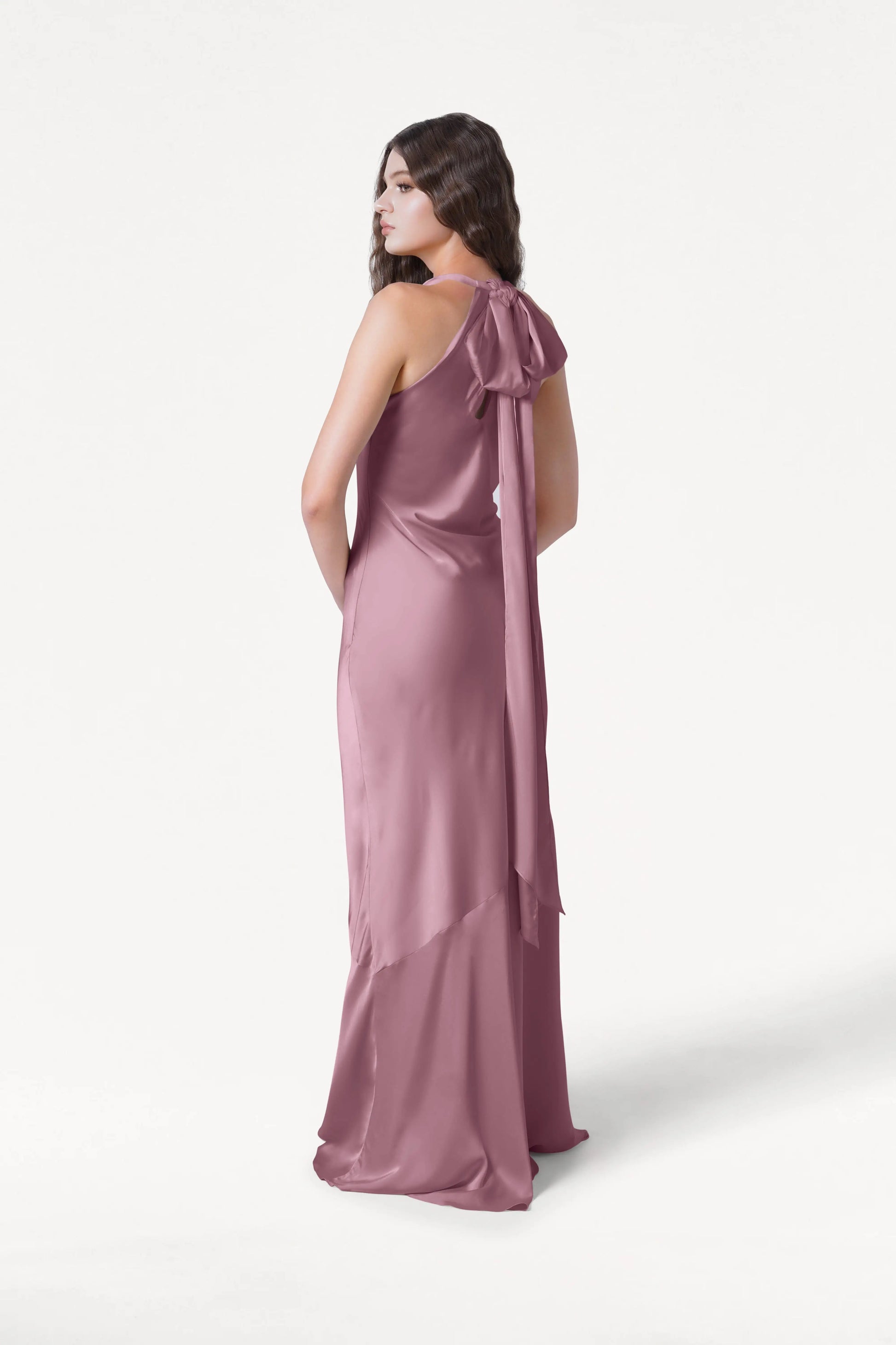 A woman with long brown hair models the Mira Bridesmaid Dress in Rose Pink Satin by TH&TH—a sleeveless, floor-length gown with a bow at the back and bias-cut silhouette—standing against a plain white background.
