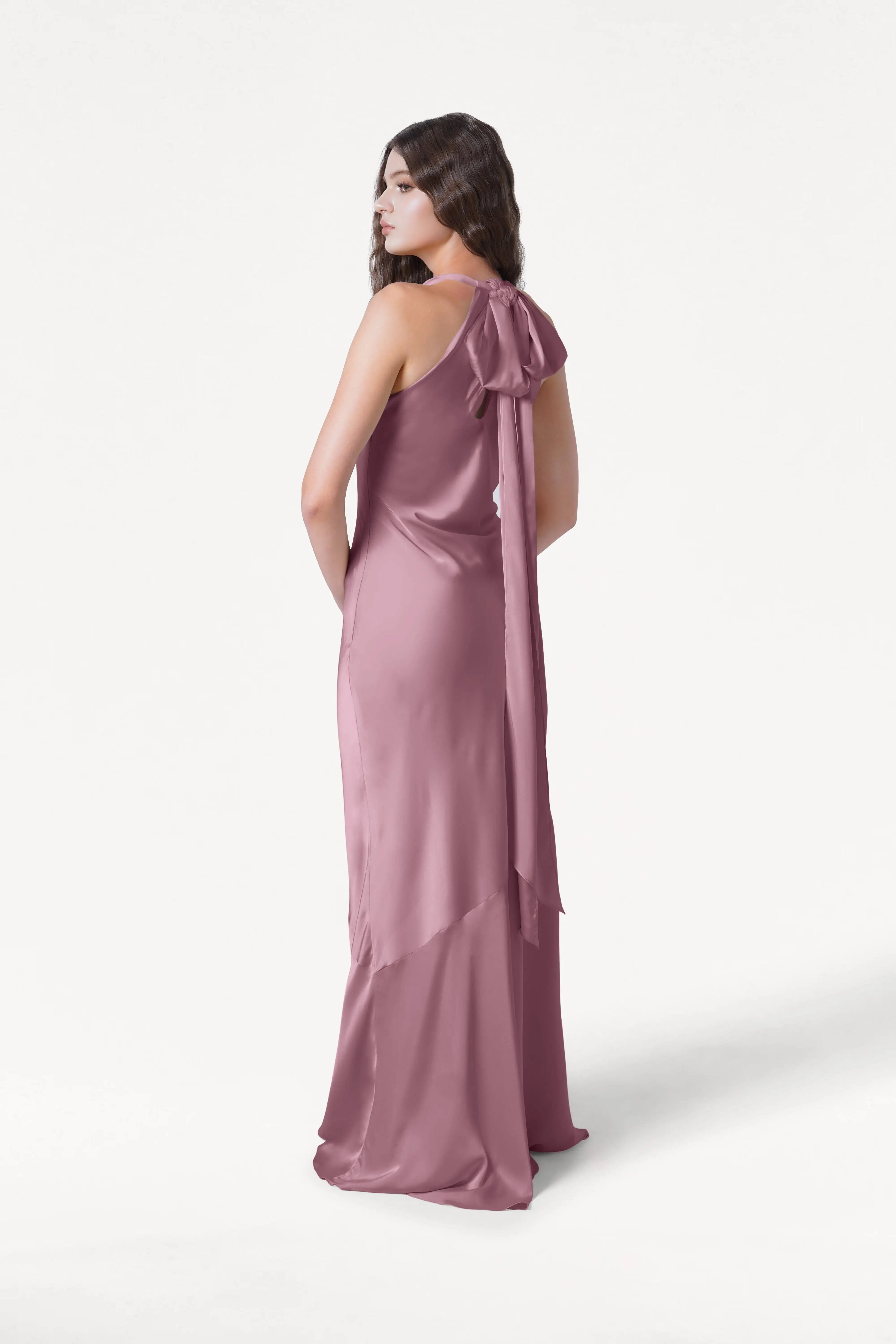 A woman with long brown hair models the Mira Bridesmaid Dress in Rose Pink Satin by TH&TH—a sleeveless, floor-length gown with a bow at the back and bias-cut silhouette—standing against a plain white background.