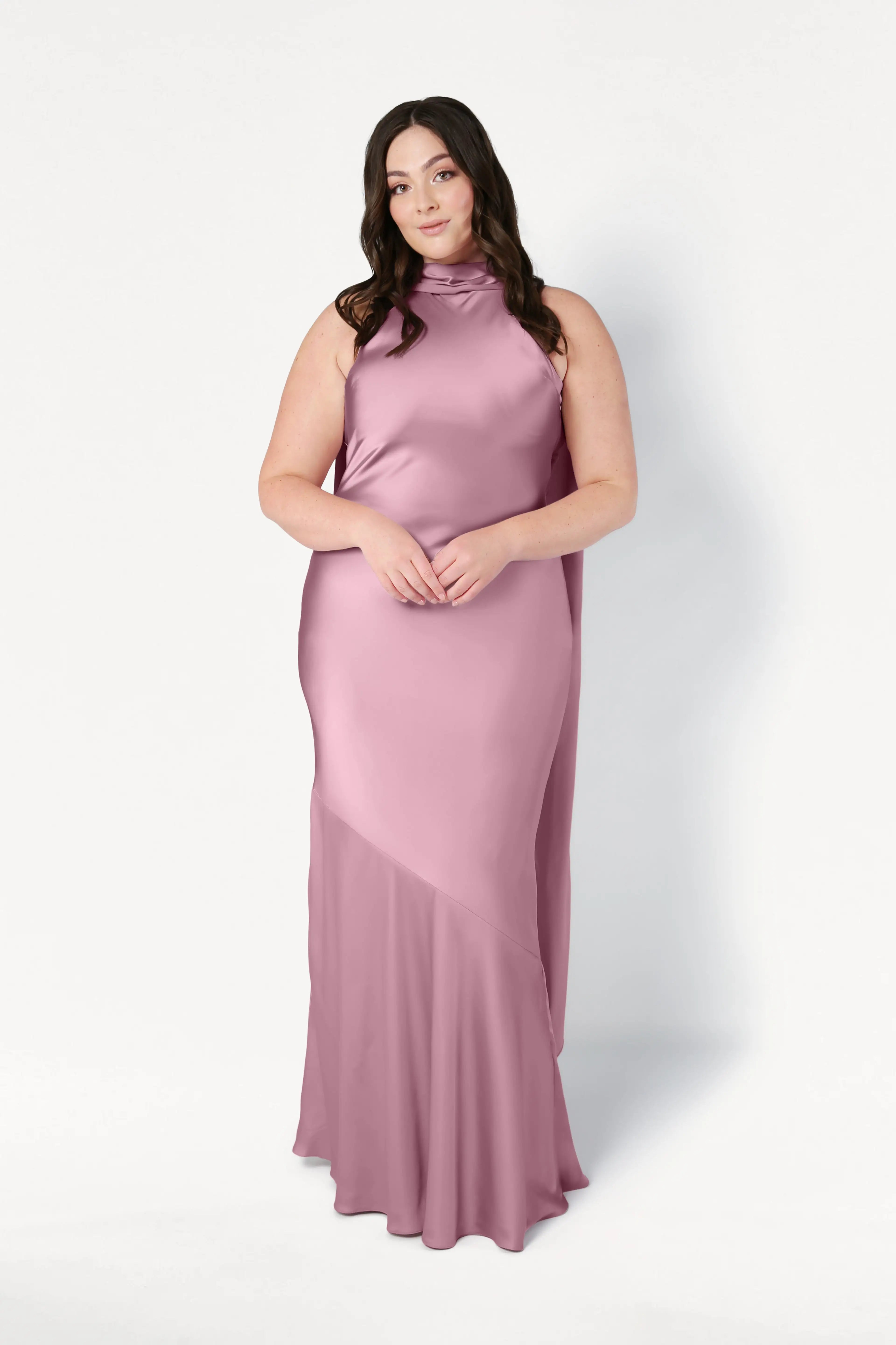 A woman with long dark hair wears the Mira Bridesmaid Dress in Rose Pink Satin by TH&TH, a sleeveless, high-neck, floor-length gown with a bias-cut silhouette. She stands confidently against a plain white background, hands clasped in front.