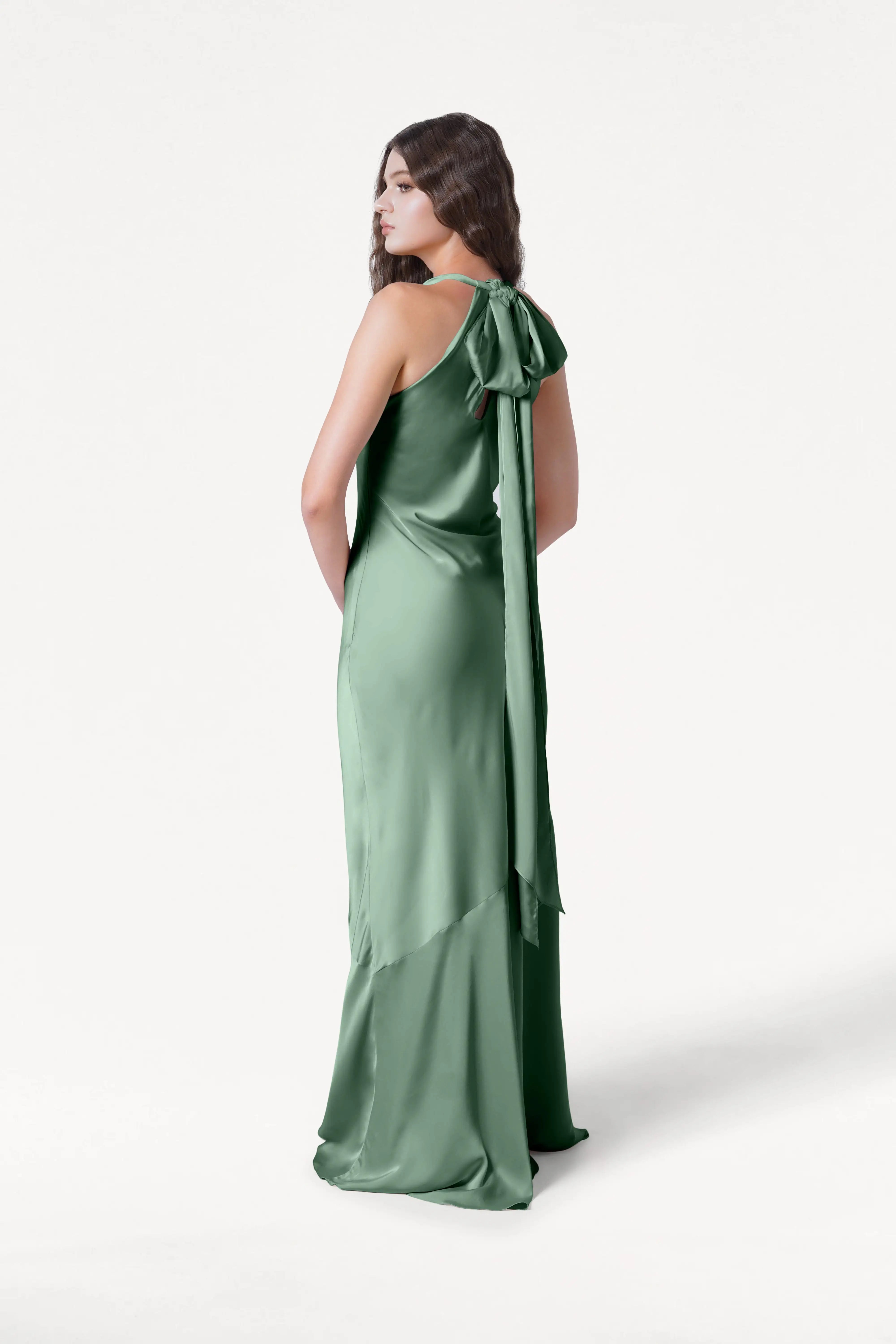 A woman faces away in the TH&TH Mira Bridesmaid Dress, crafted from sage green satin with a large back bow and bias-cut silhouette. Her dark wavy hair cascades over her shoulders against a plain white background.