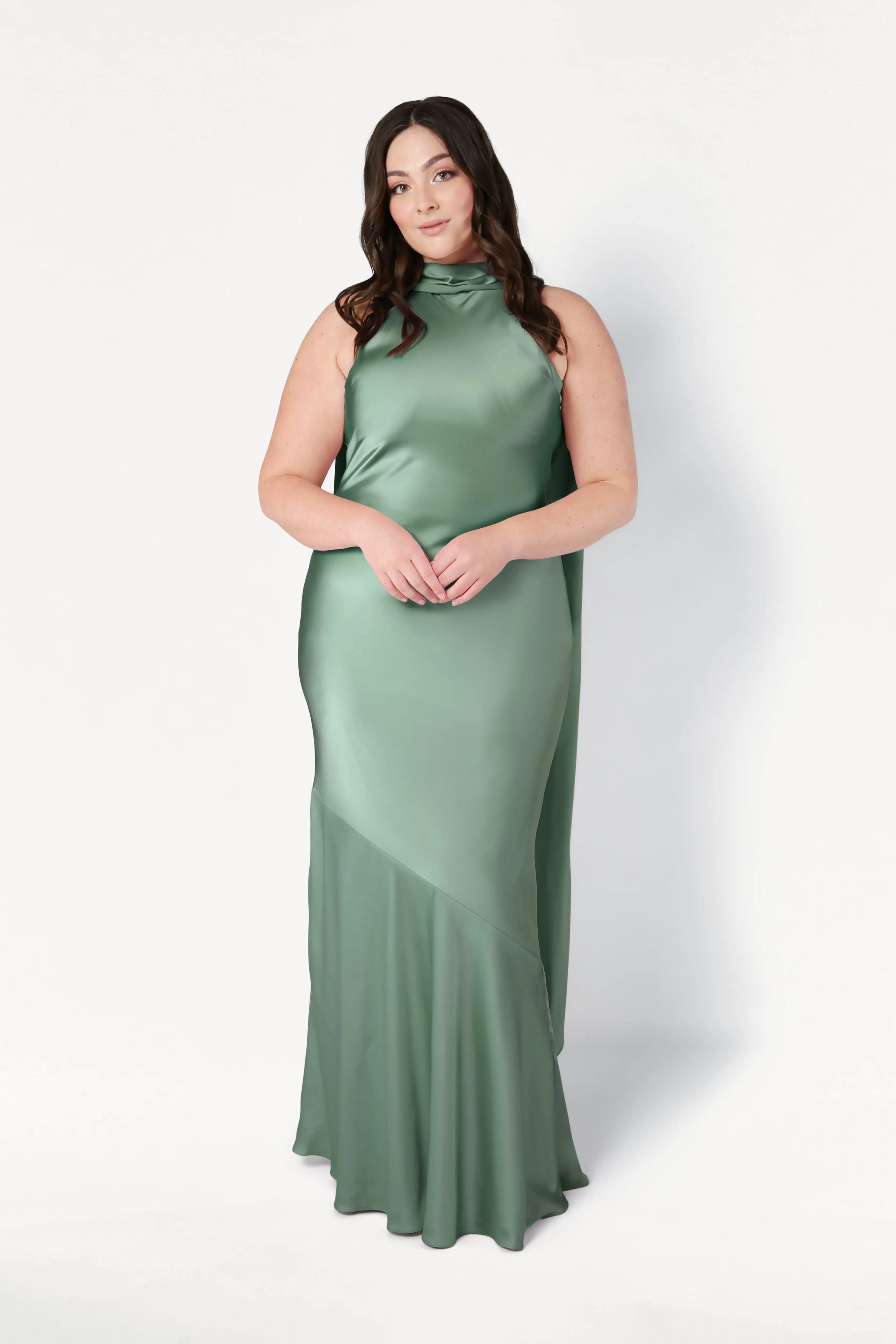 A woman with long dark hair wears the TH&TH Mira Bridesmaid Dress in sage green satin, a sleeveless, floor-length gown with a high neckline, standing against a plain white background.