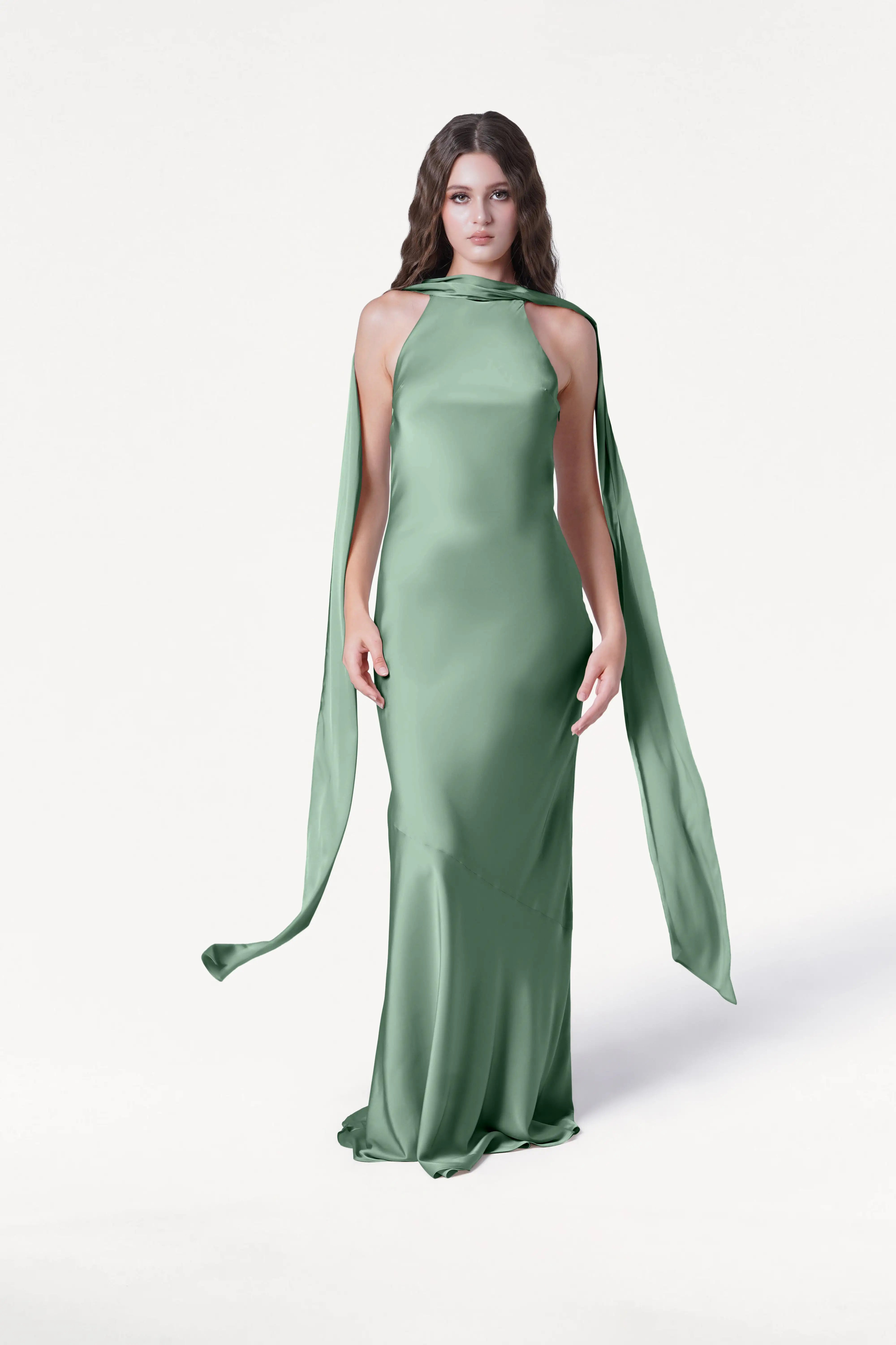 A woman wears the TH&TH Mira Bridesmaid Dress in sage green satin, featuring a high neckline and long, draped panels, standing against a plain white background.