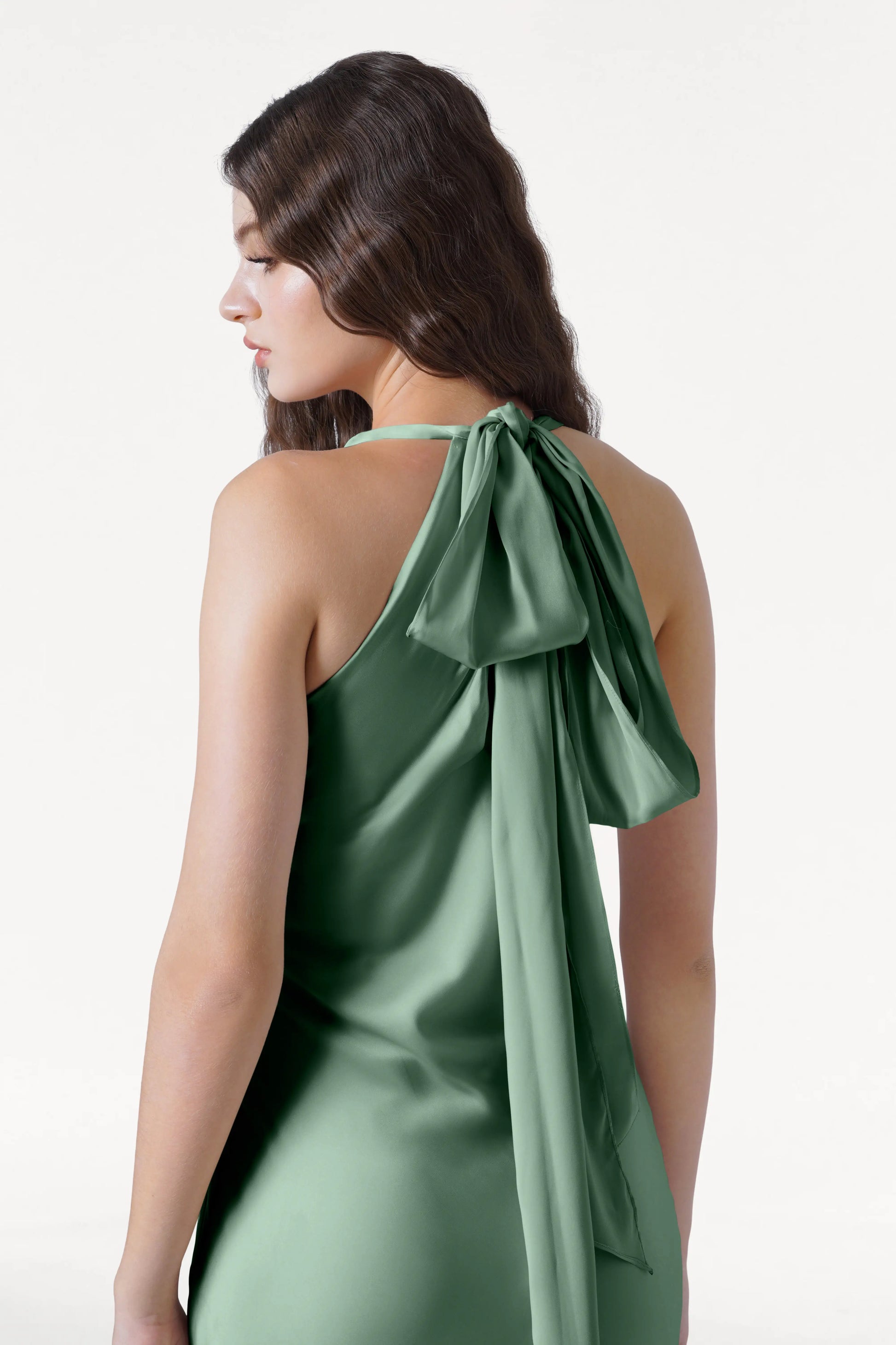 A woman with long, wavy brown hair models the TH&TH Mira Bridesmaid Dress in sage green satin, featuring a halterneck and large back bow, standing against a plain white background.