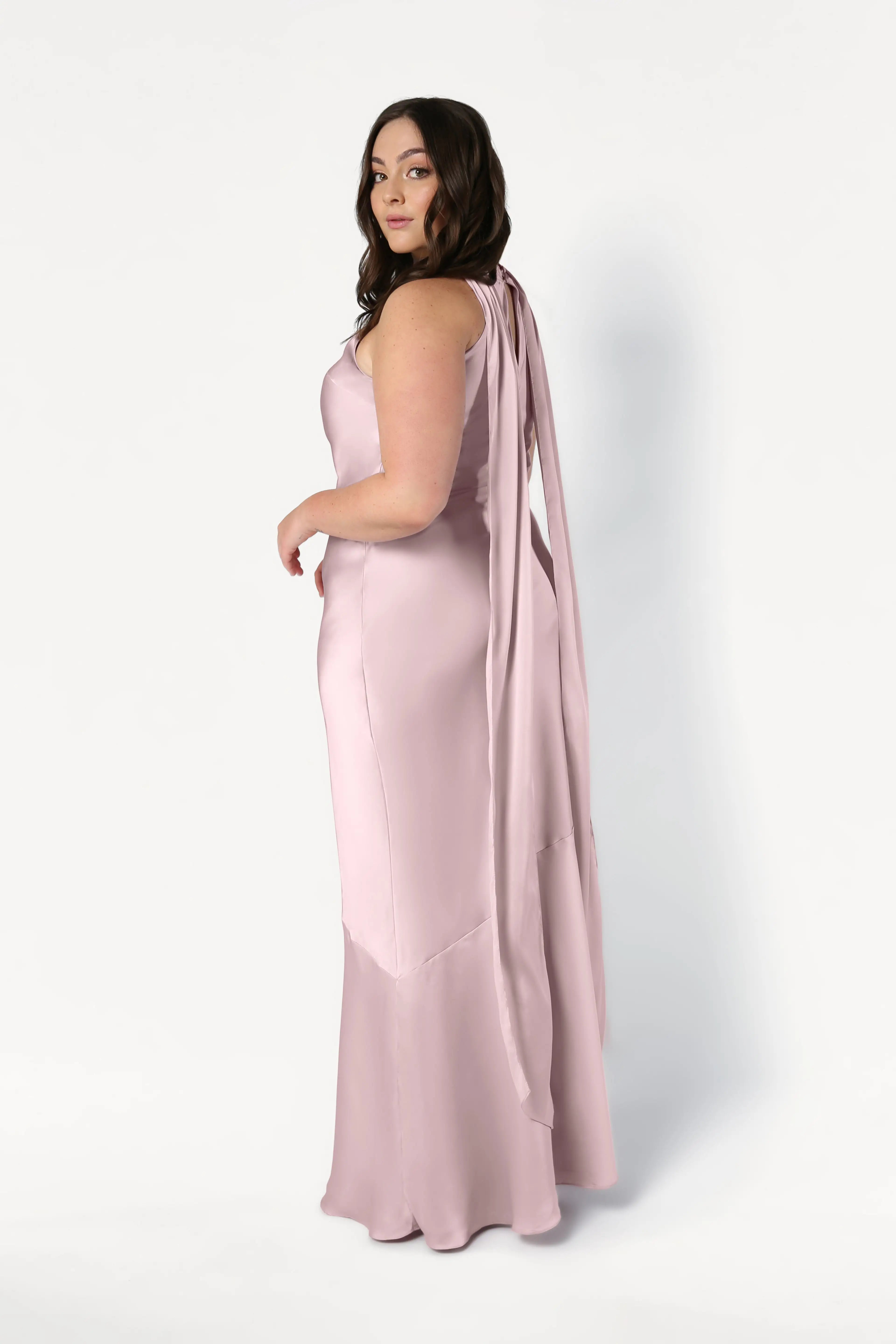 A woman in the TH&TH Mira Bridesmaid Dress in Smoked Blush Satin stands sideways against a white background, looking over her shoulder and softly smiling, showcasing the dress’s elegant draped tie back.