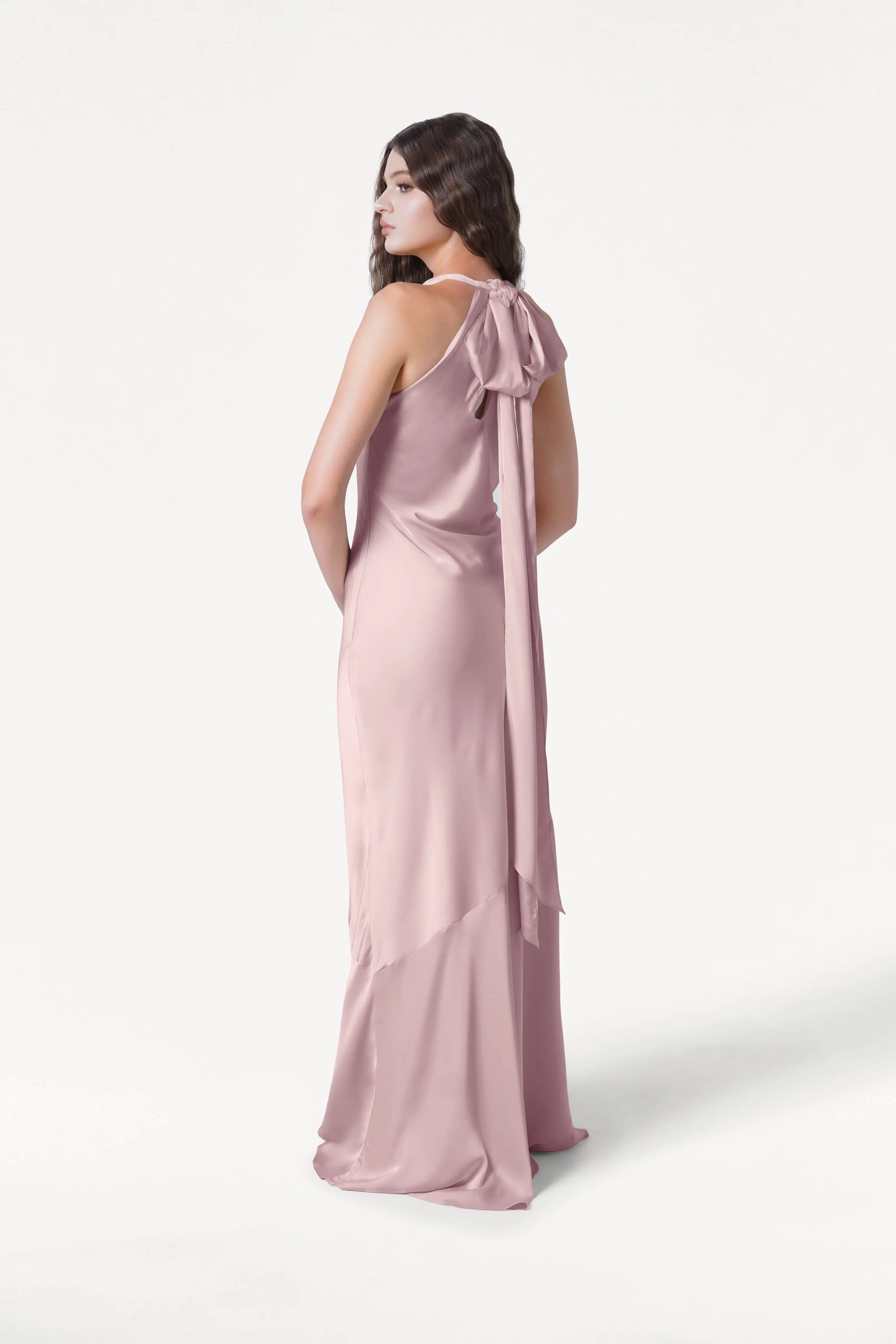 A woman stands turned slightly away in the TH&TH Mira Bridesmaid Dress in Smoked Blush Satin, featuring a large back neck bow, sleeveless design, and a soft flowing hem, set against a plain white background.