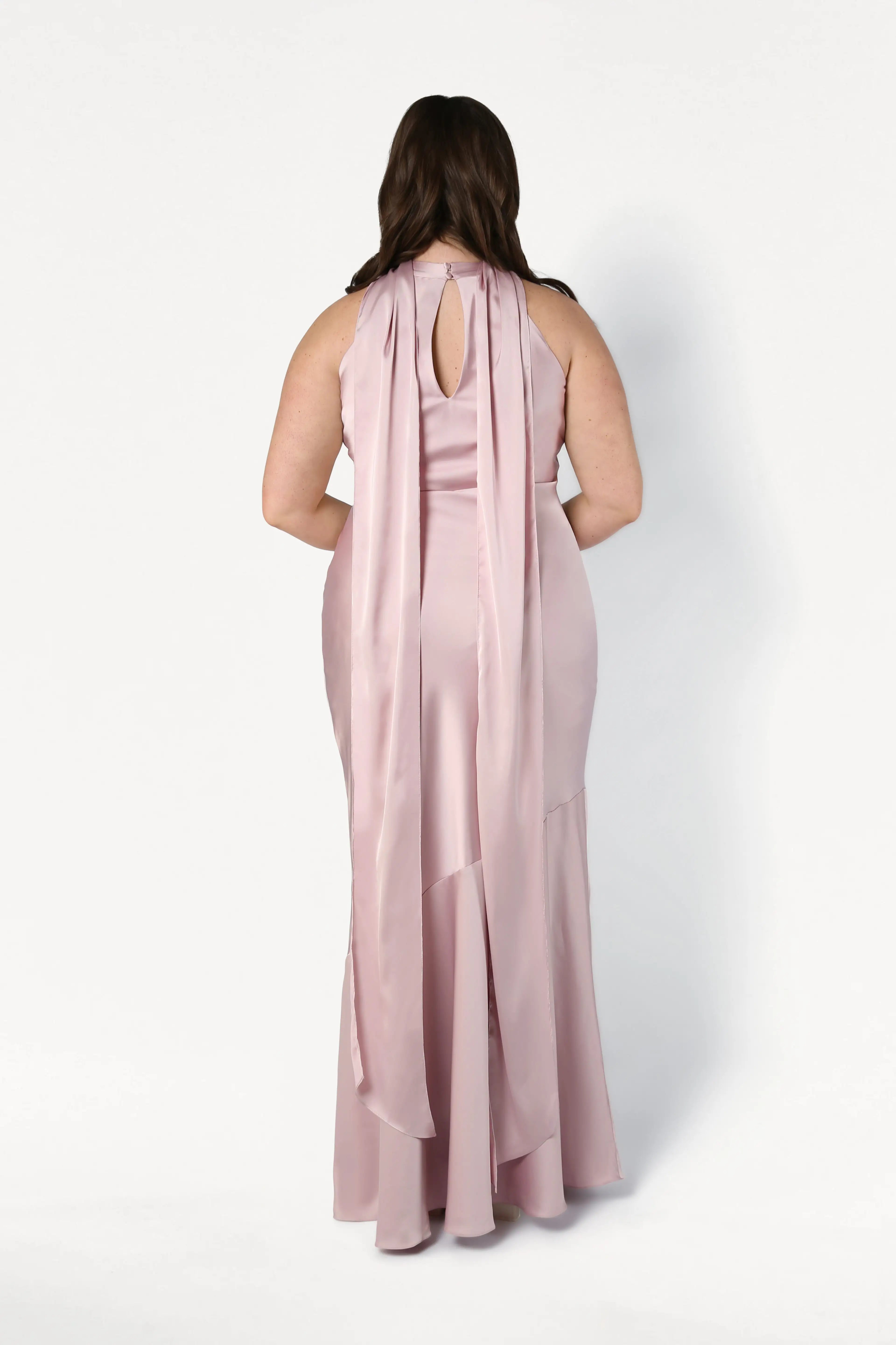A woman with long brown hair wears the TH&TH Mira Bridesmaid Dress in Smoked Blush Satin, a floor-length, sleeveless gown with a keyhole back and flowing panels, standing against a plain white background, facing away.