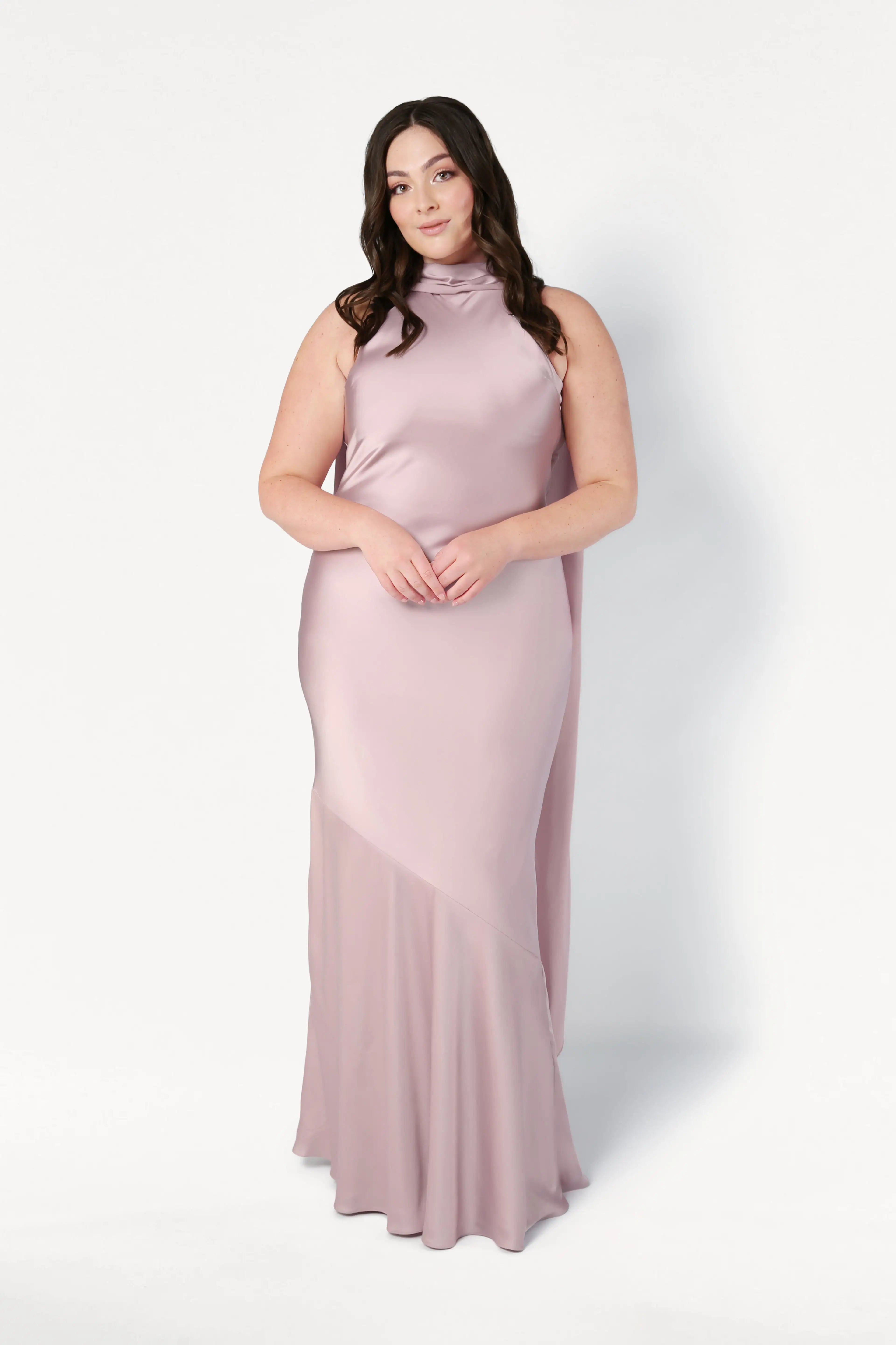 A woman with long, wavy brown hair wears the TH&TH Mira Bridesmaid Dress in smoked blush satin, featuring a floor-length, bias-cut silhouette and high neckline, standing against a plain white background.