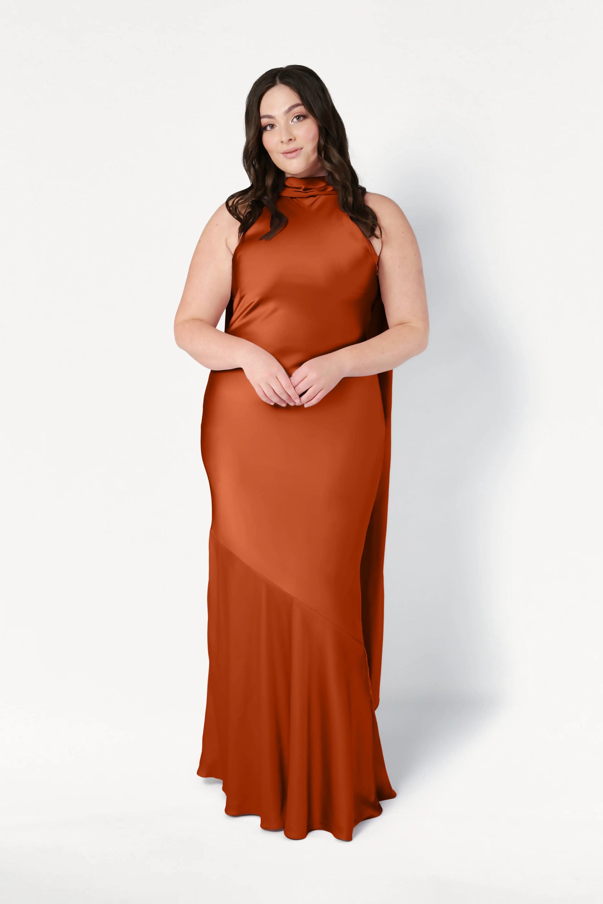 A woman with long dark hair stands against a plain white background, hands gently clasped, wearing the TH&TH Mira Bridesmaid Dress in Terracotta Satin—a sleeveless, floor-length gown.