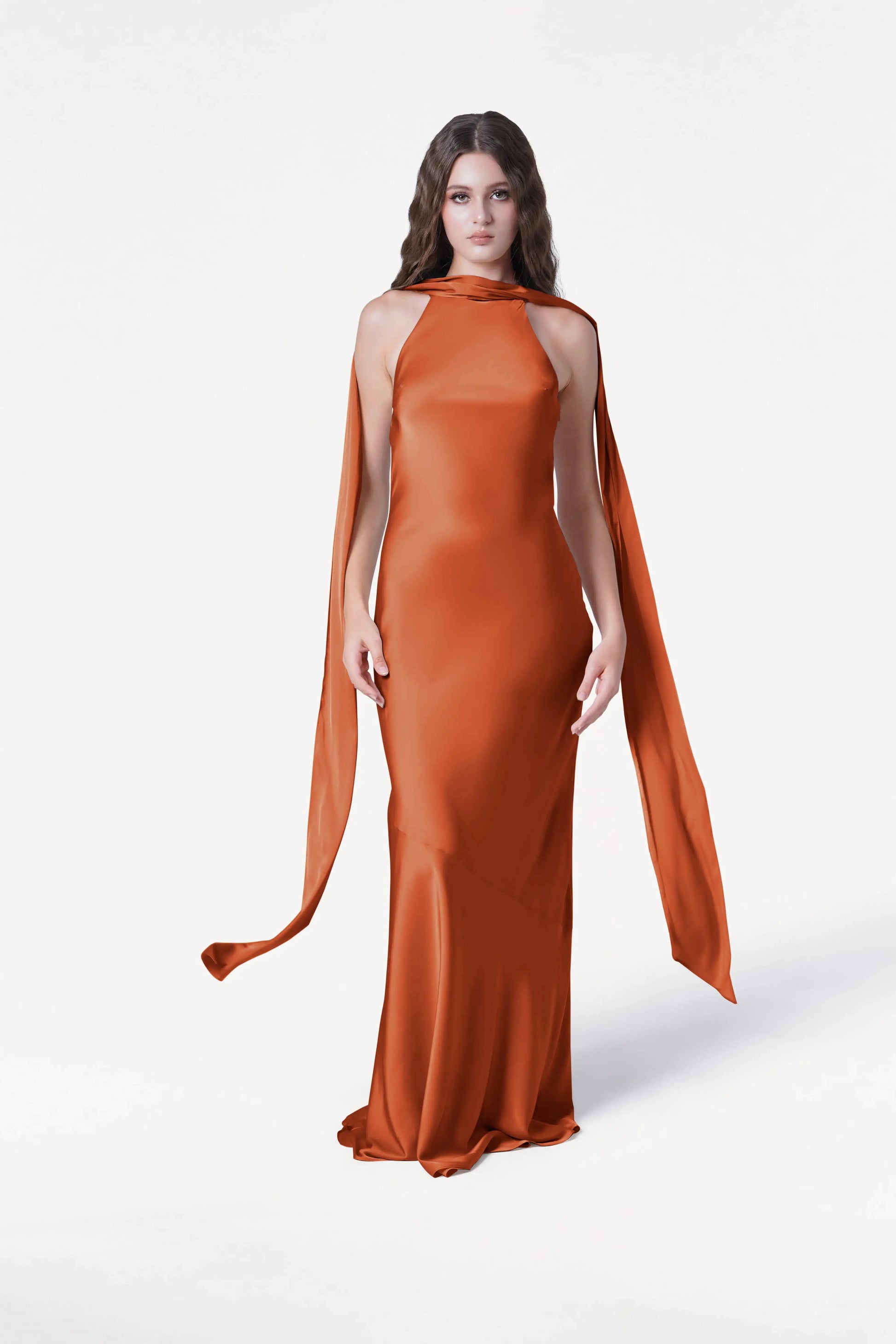 A woman with long brown hair models the TH&TH Mira Bridesmaid Dress in Terracotta Satin, a floor-length gown with a high neckline and flowing shoulder scarves, standing against a plain white background.
