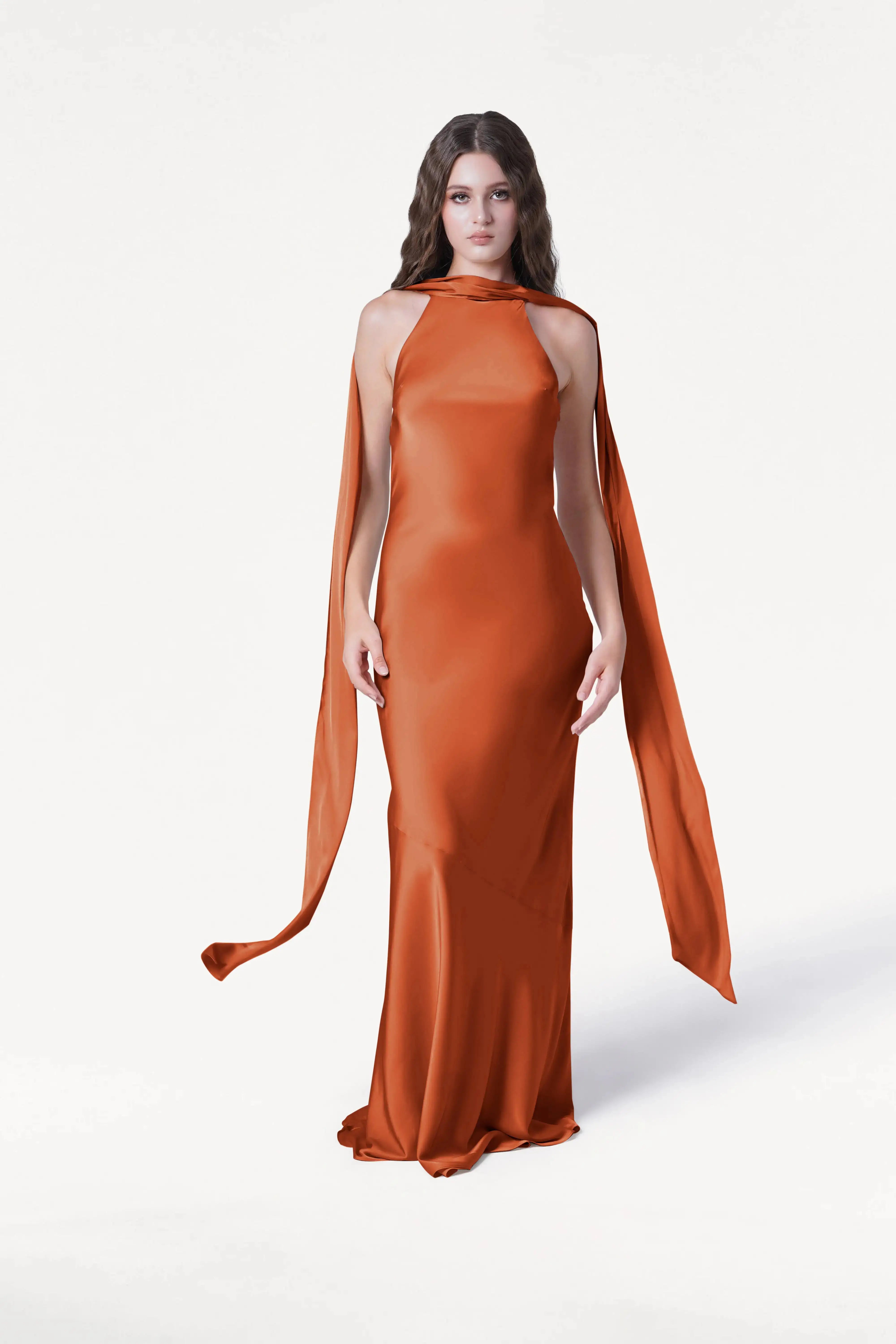 A woman with long brown hair models the TH&TH Mira Bridesmaid Dress in Terracotta Satin, a floor-length gown with a high neckline and flowing shoulder scarves, standing against a plain white background.