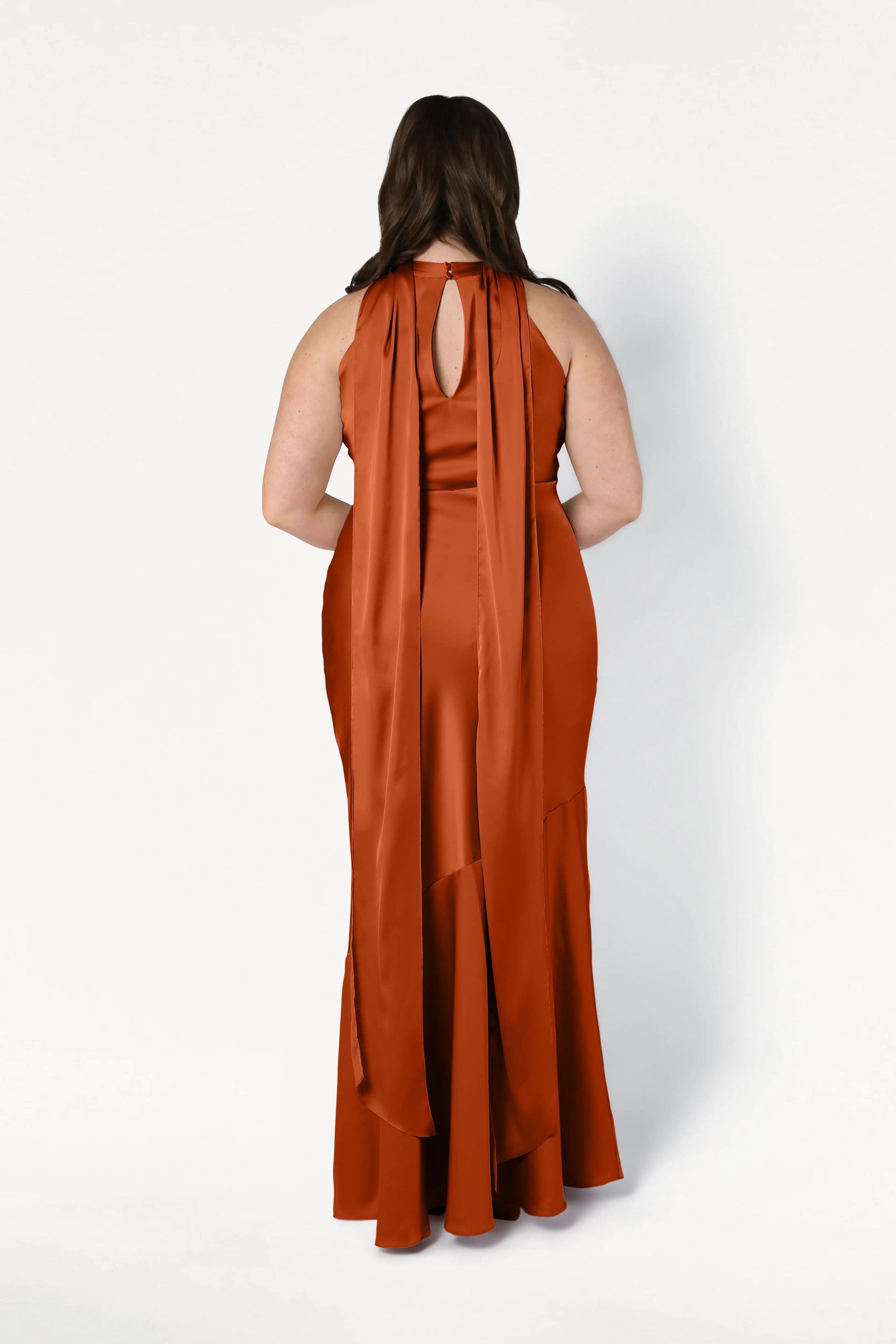 A woman faces away, wearing the TH&TH Mira Bridesmaid Dress in Terracotta Satin, which features two wide sashes draping down her back, set against a plain white backdrop.