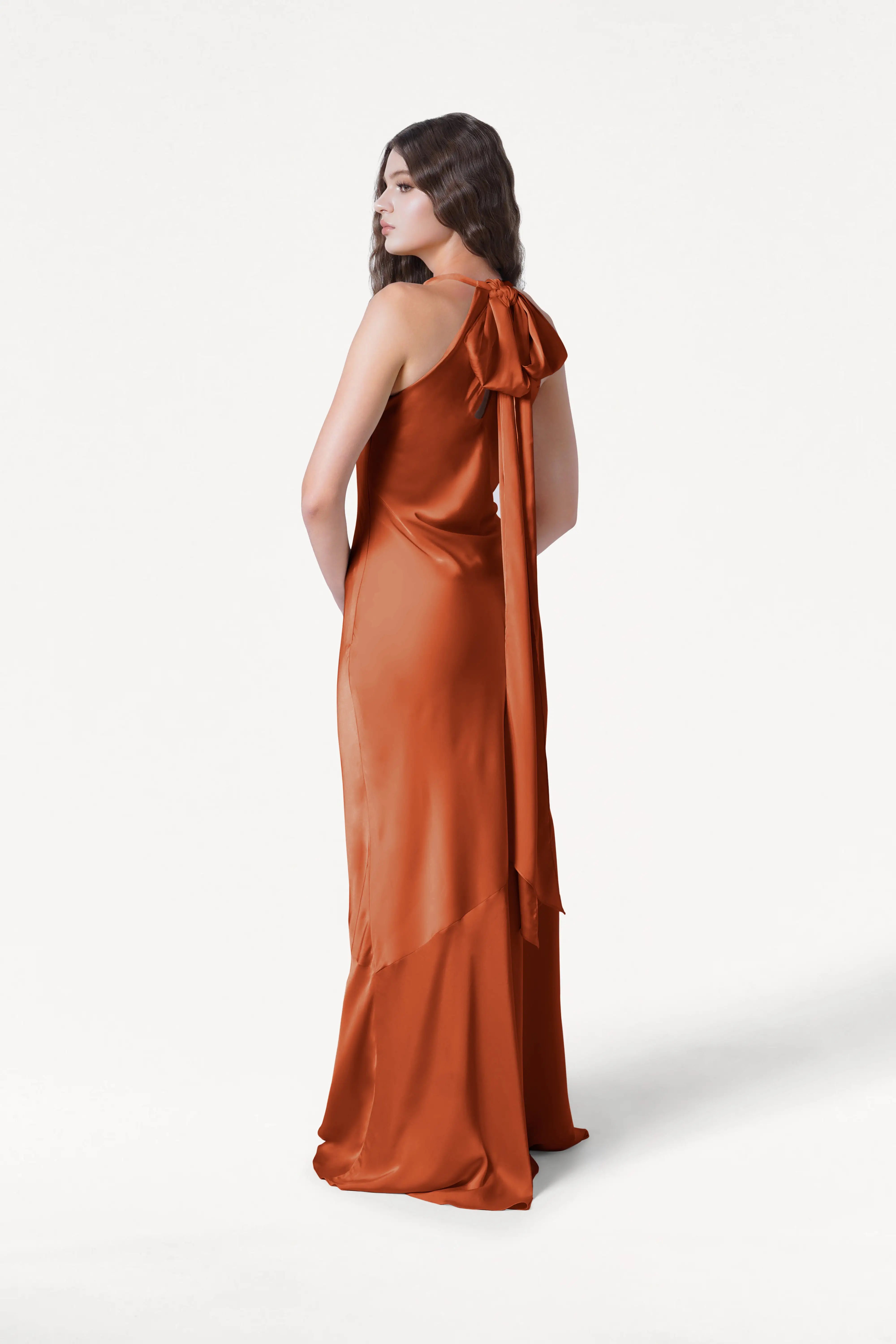 A woman wears the TH&TH Mira Bridesmaid Dress in Terracotta Satin, featuring a long, sleeveless design with a back-neck bow and flowing skirt. She stands against a plain white background, facing away and looking over her shoulder.