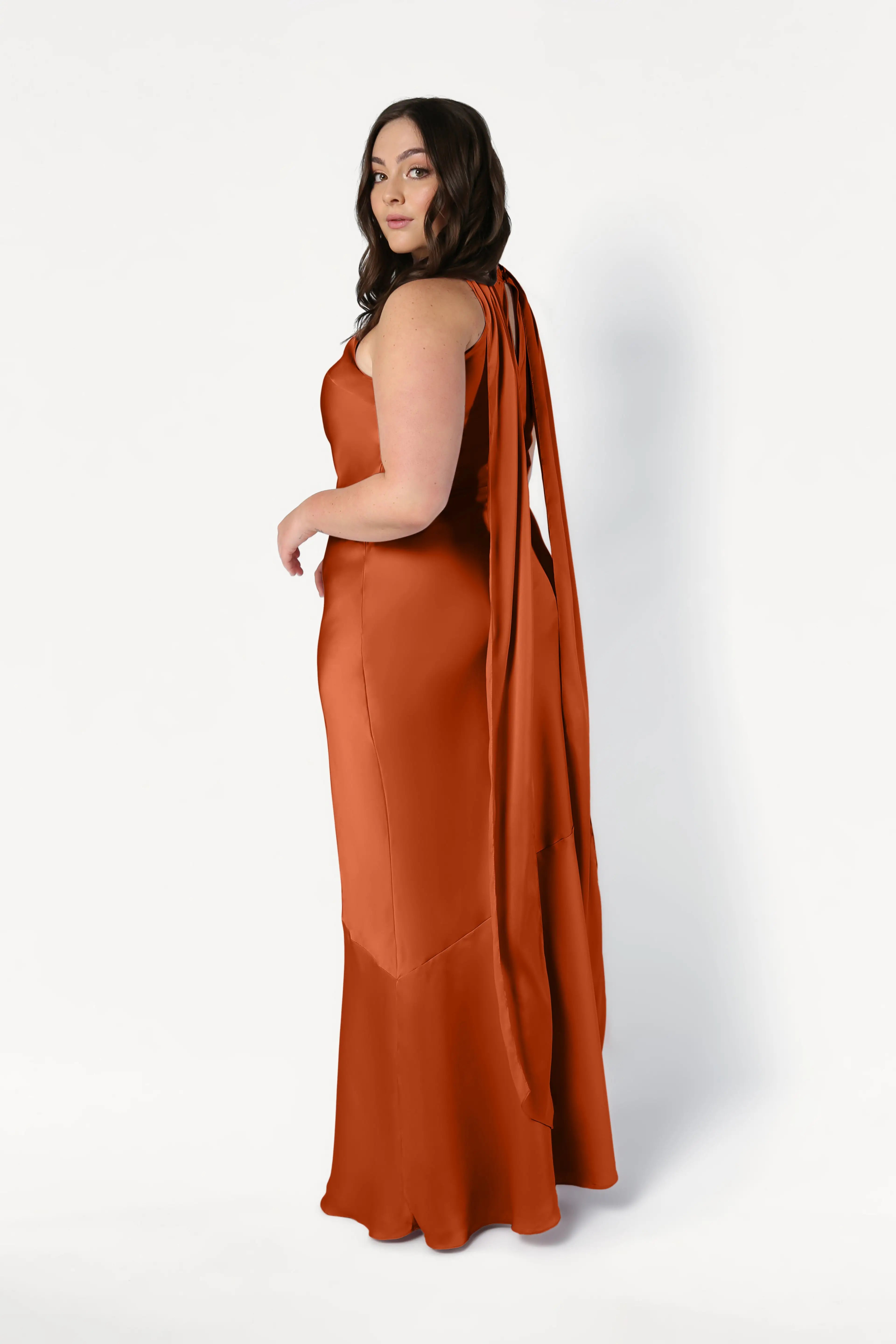 A woman in the TH&TH Mira Bridesmaid Dress in Terracotta Satin stands against a white background, looking over her shoulder. The floor-length dress features long, flowing tie details at the back.