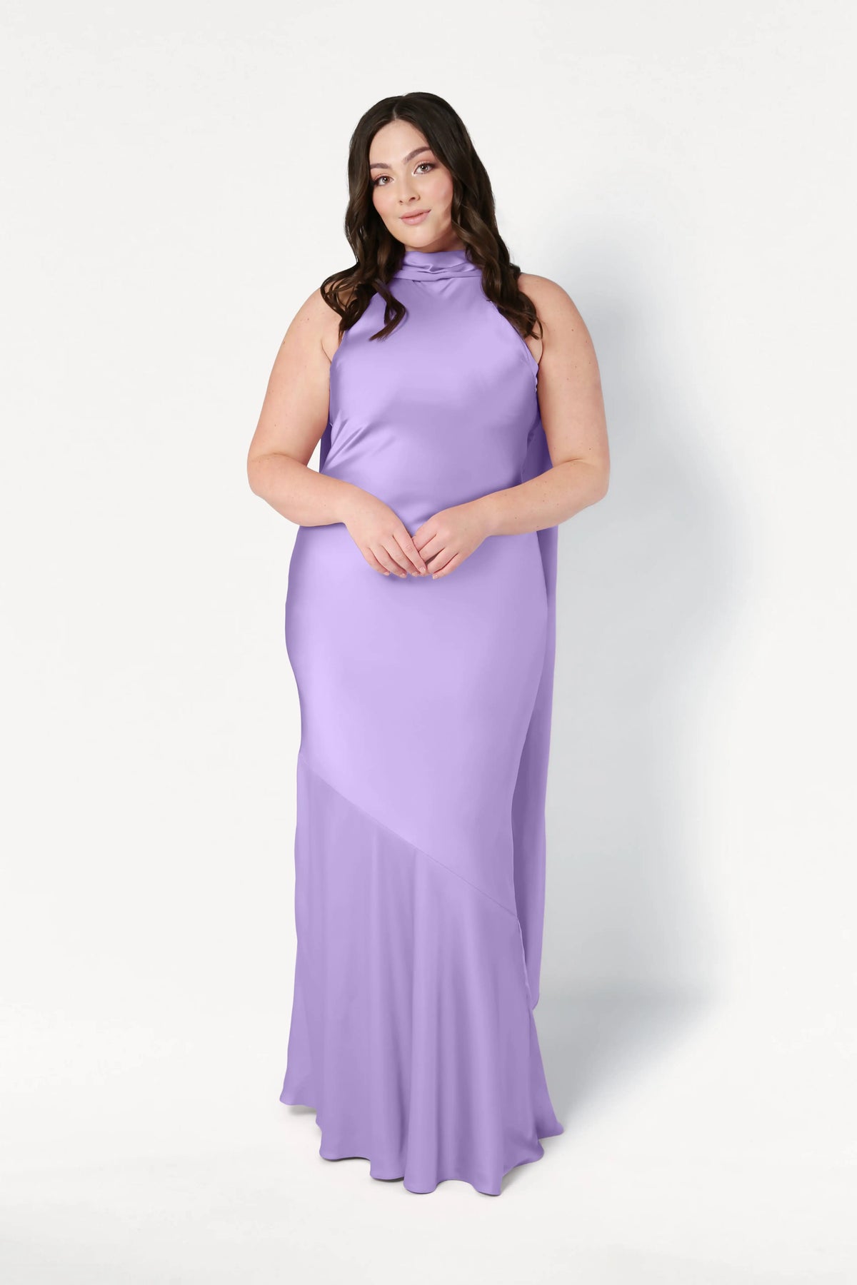 A woman with long dark hair stands against a white background wearing the TH&TH Mira Bridesmaid Dress in floor-length violet satin, sleeveless with a high neckline. She faces forward, hands gently clasped in front of her.