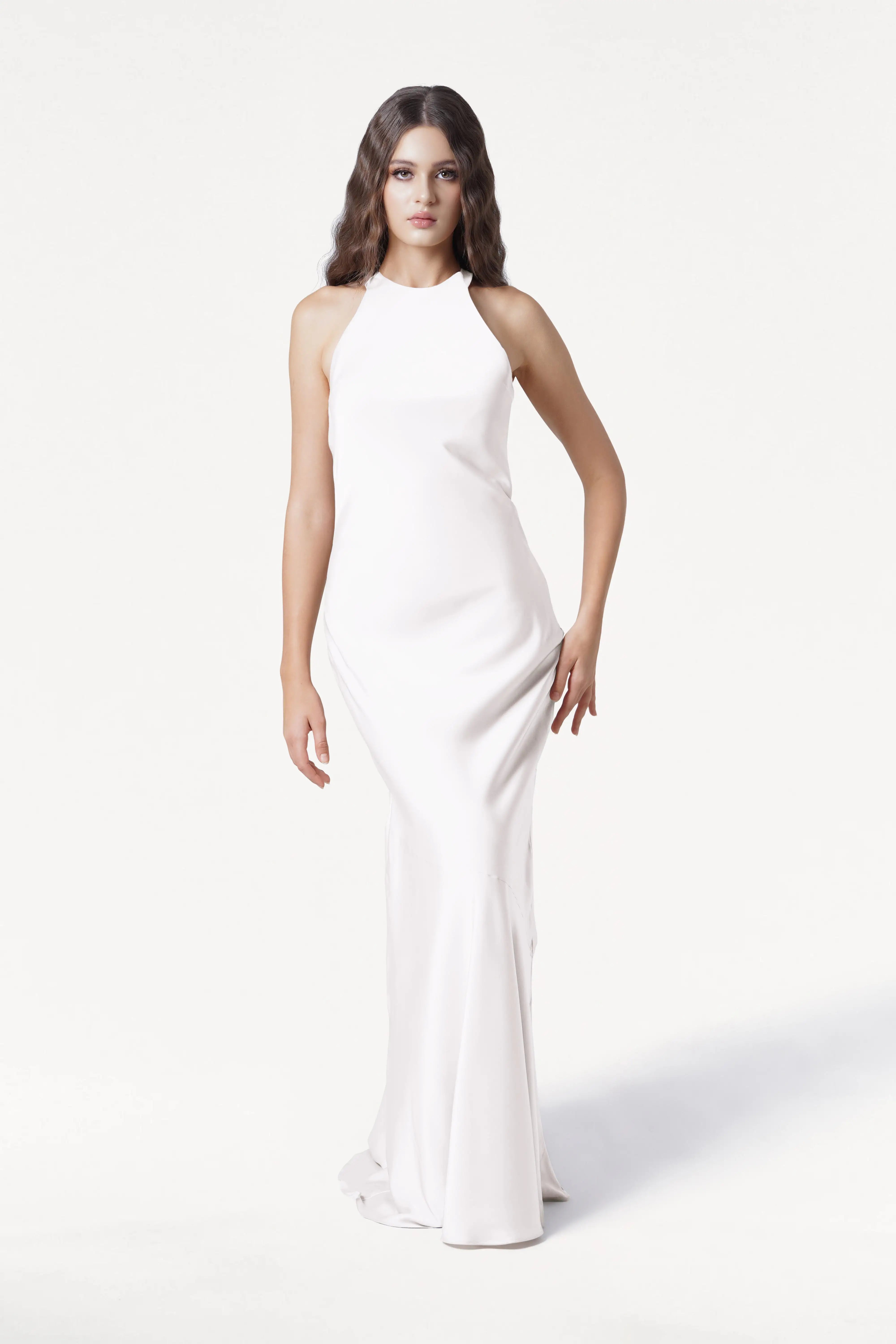 A woman with long wavy brown hair models the sleeveless, floor-length Mira Dress in Ivory Satin by TH&TH, featuring a high neckline, against a plain white background.