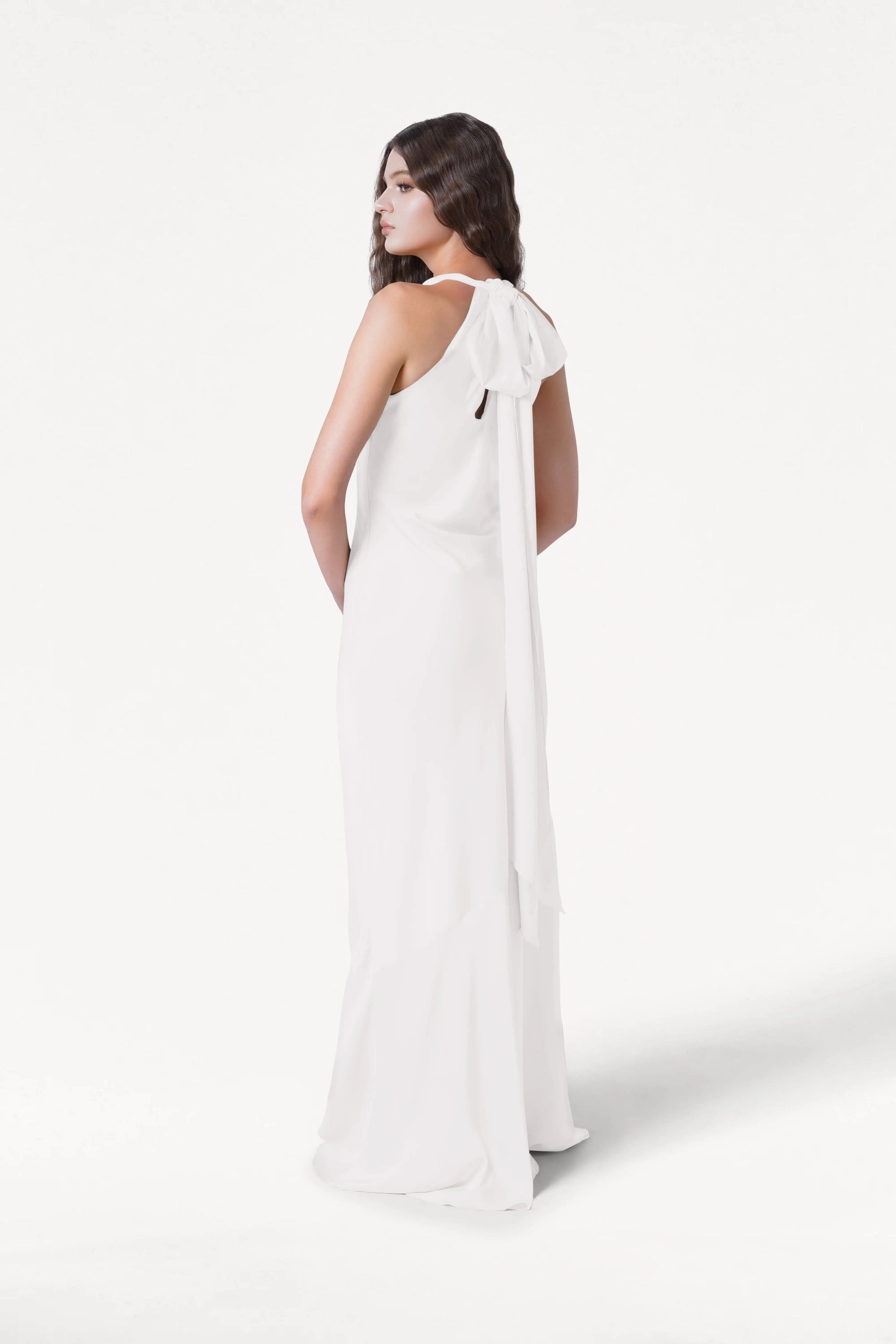 A woman in the TH&TH Mira Dress in Ivory Satin stands facing away, looking over her shoulder. The flowing white gown features a halterneck with a large bow and an elegant silhouette against a white background.