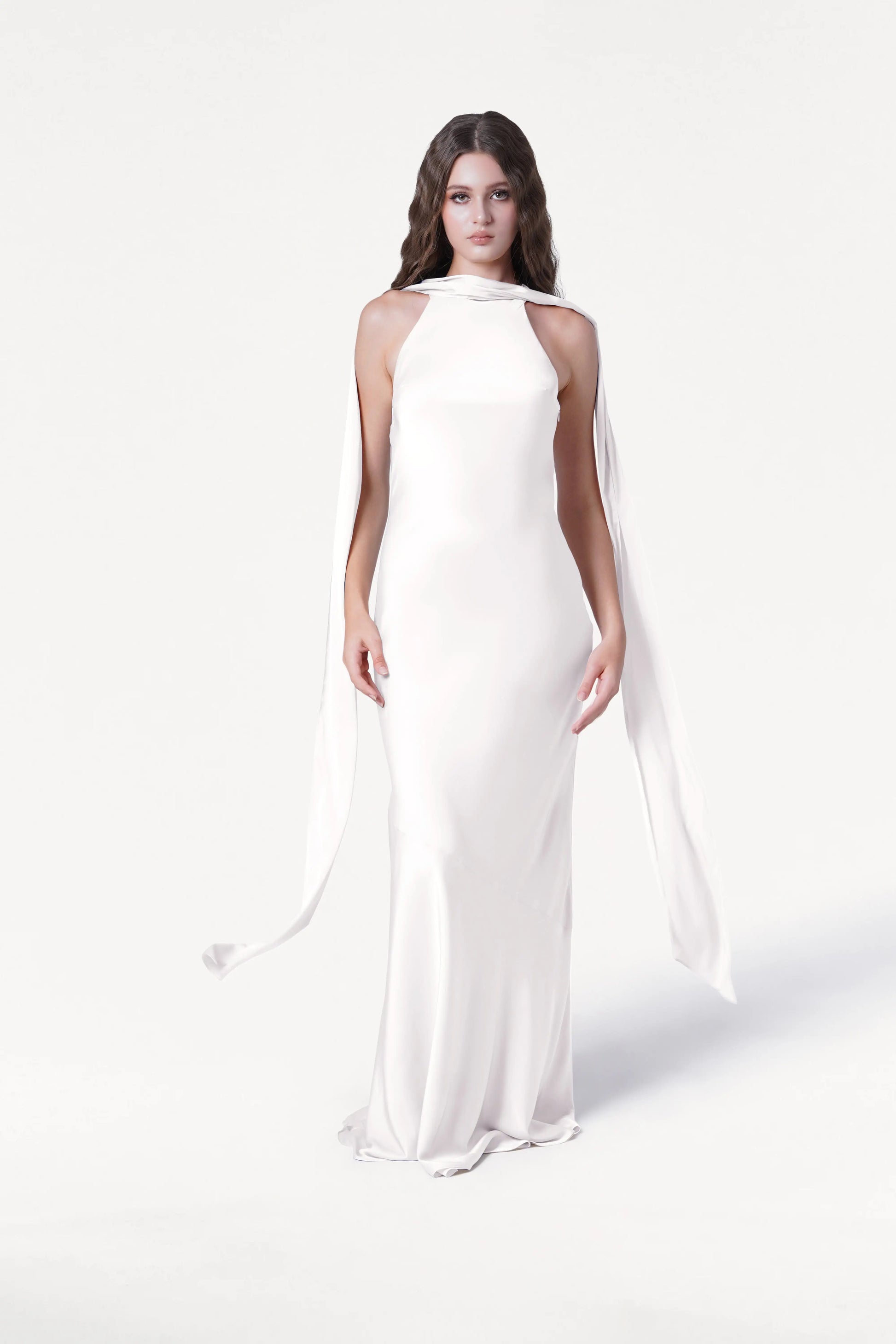 A person with long wavy hair models the TH&TH Mira Dress in Ivory Satin, a sleek floor-length bridesmaid gown featuring draped shoulder fabric, standing against a plain white background.