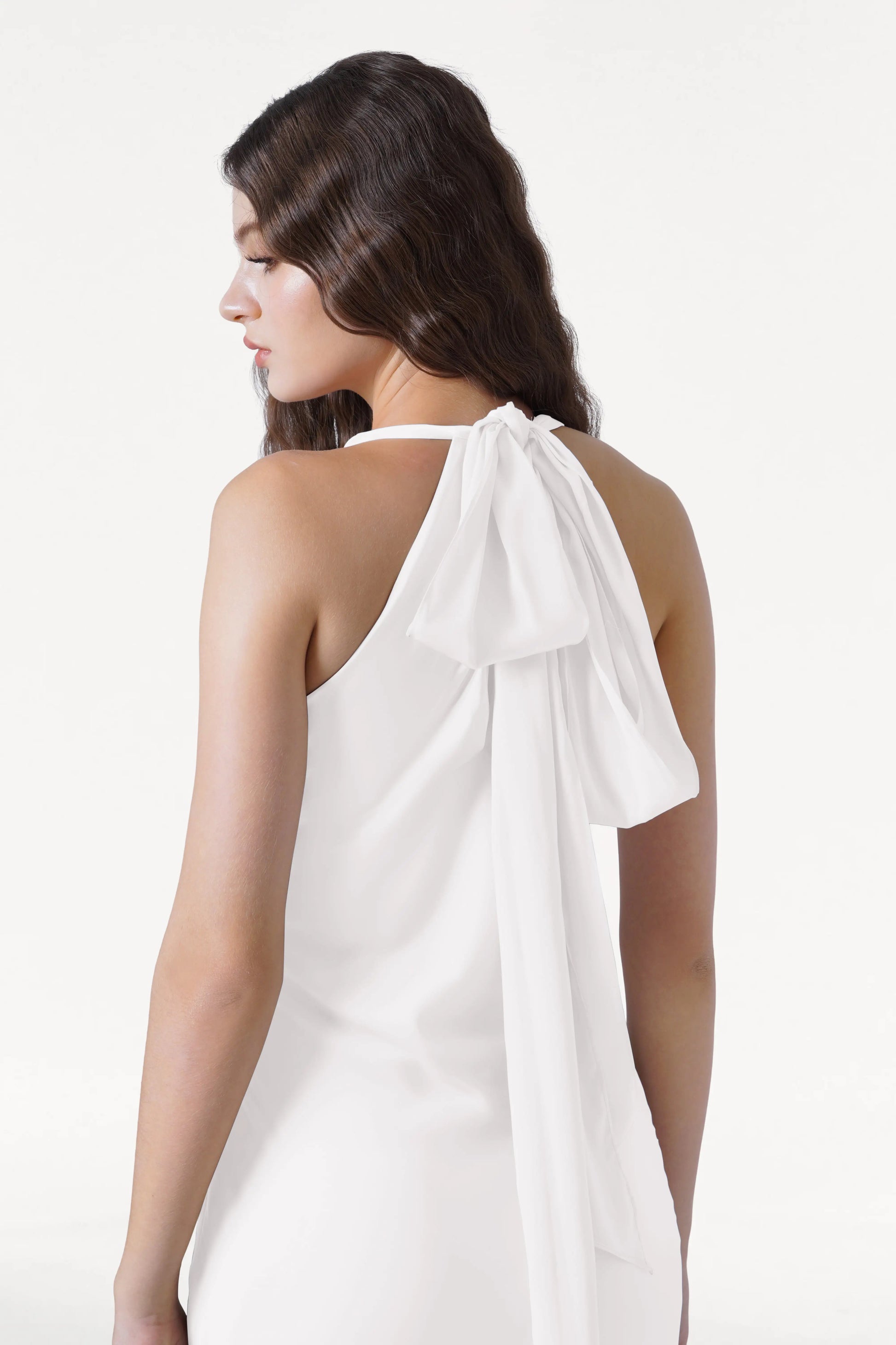 A woman with long, wavy brown hair models the Mira Dress in Ivory Satin by TH&TH, a sleeveless bridesmaid gown with a large bow and flowing sash at the back, standing against a plain white background.