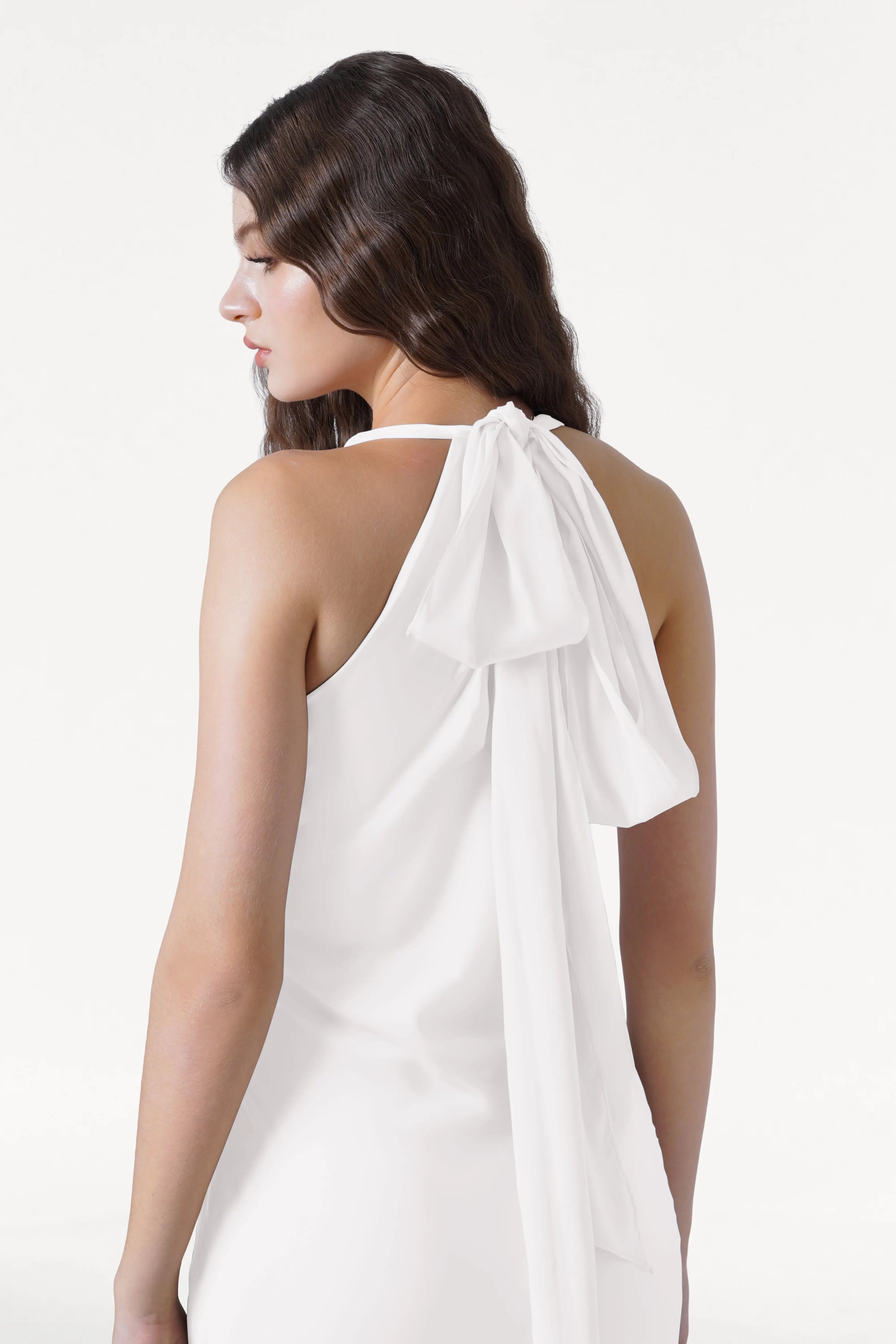 A woman with long, wavy brown hair models the Mira Dress in Ivory Satin by TH&TH, a sleeveless bridesmaid gown with a large bow and flowing sash at the back, standing against a plain white background.