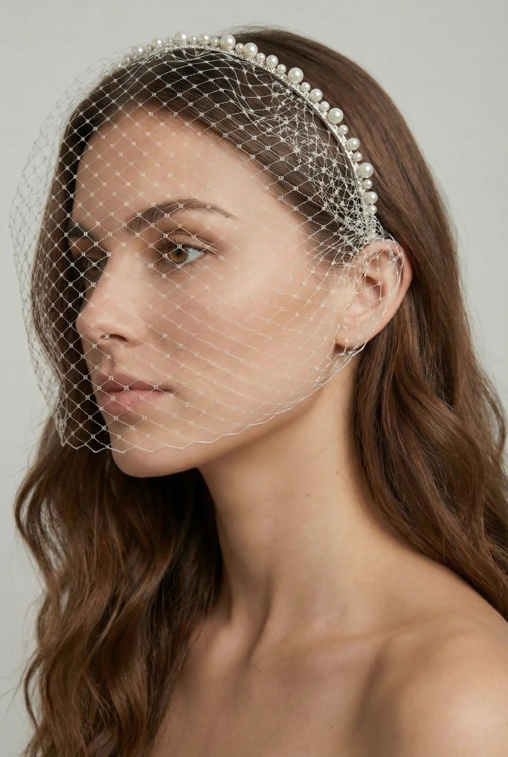A woman with long brown hair wears the TH&TH Moreau Headband with Birdcage, featuring a pearl design and delicate veil that partly covers her face. She has a neutral expression and stands before a plain background.