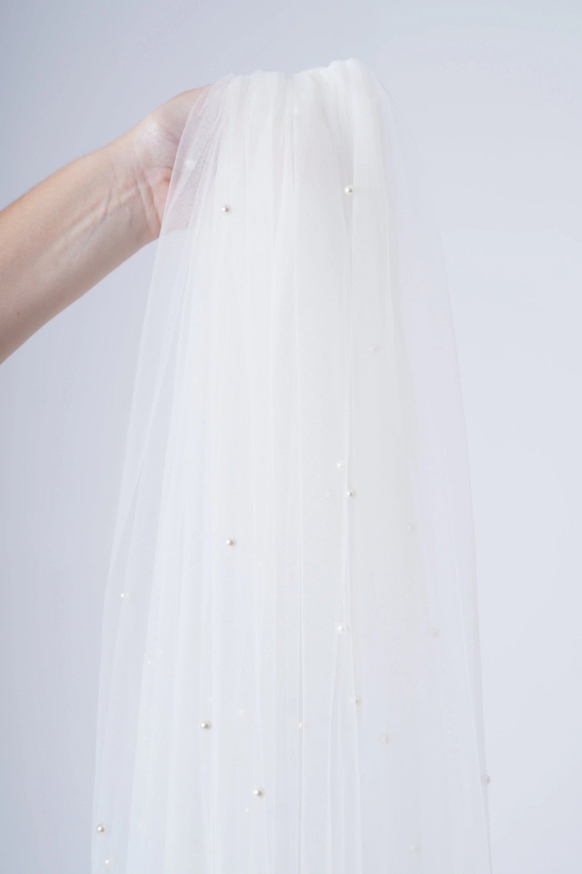 A hand displays the Muse Pearl Scatter Veil by TH&TH—a sheer white tulle veil adorned with delicate pearl-like beads—set against a plain light background.