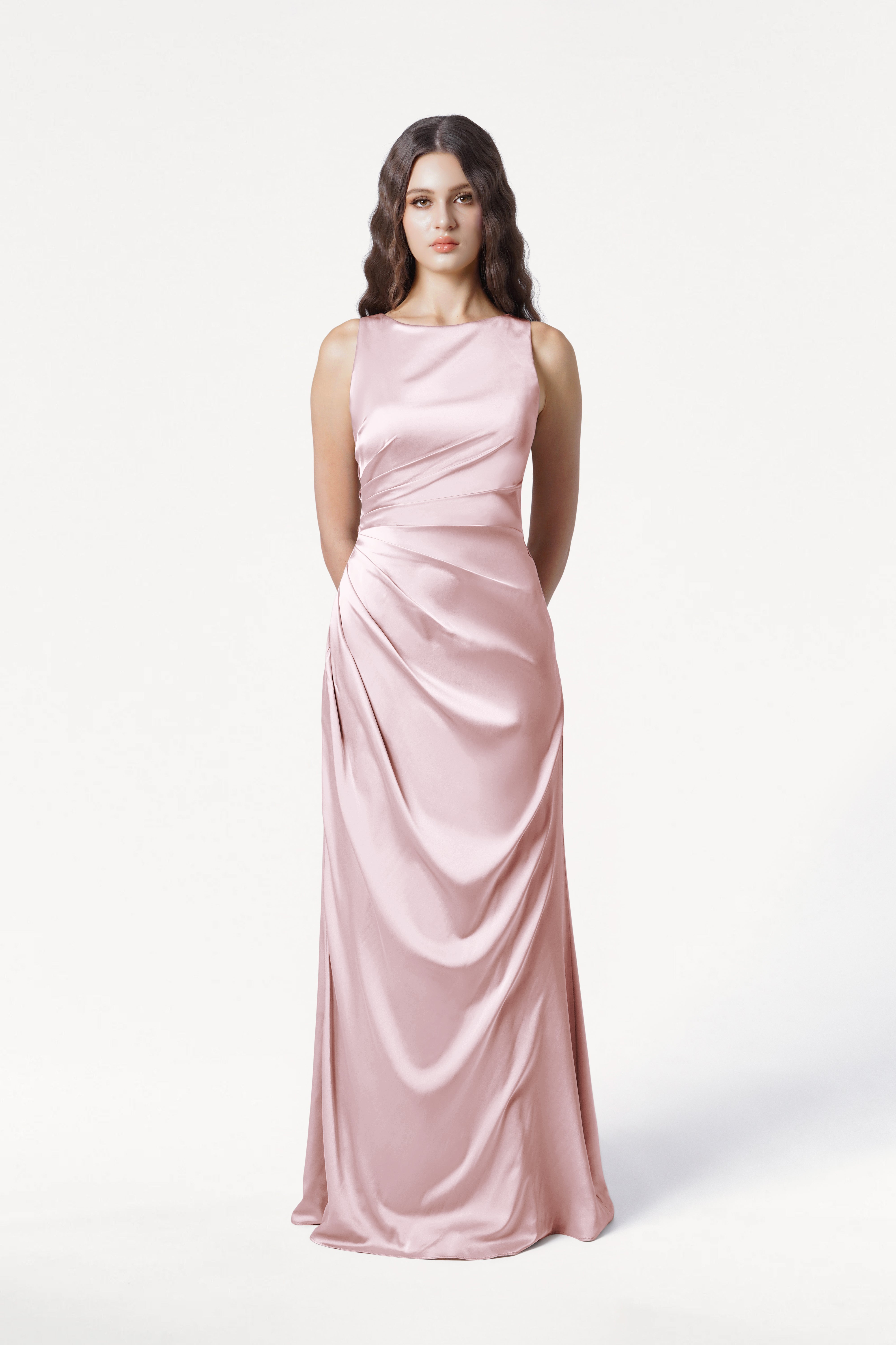 A woman with long wavy brown hair wears the TH&TH Nala Bridesmaid Dress in Blush Pink Satin, featuring Grecian-inspired pleating at the waist, sleeveless and floor-length, standing against a plain white background.