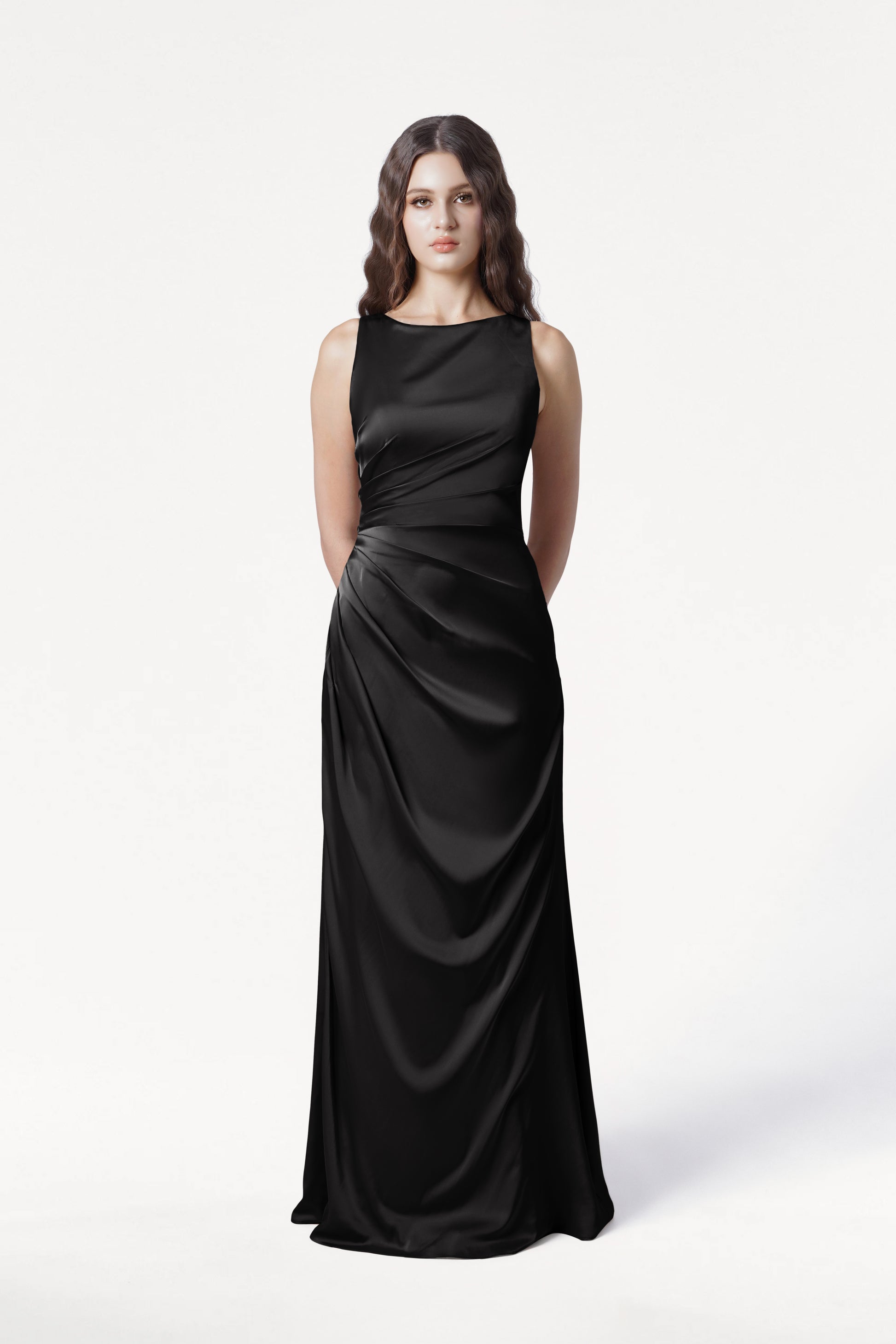 A woman with long wavy brown hair models the size-inclusive Nala Bridesmaid Dress in Black Satin by TH&TH, a floor-length gown with Grecian-inspired pleating. She stands with her hands behind her back against a plain white background.
