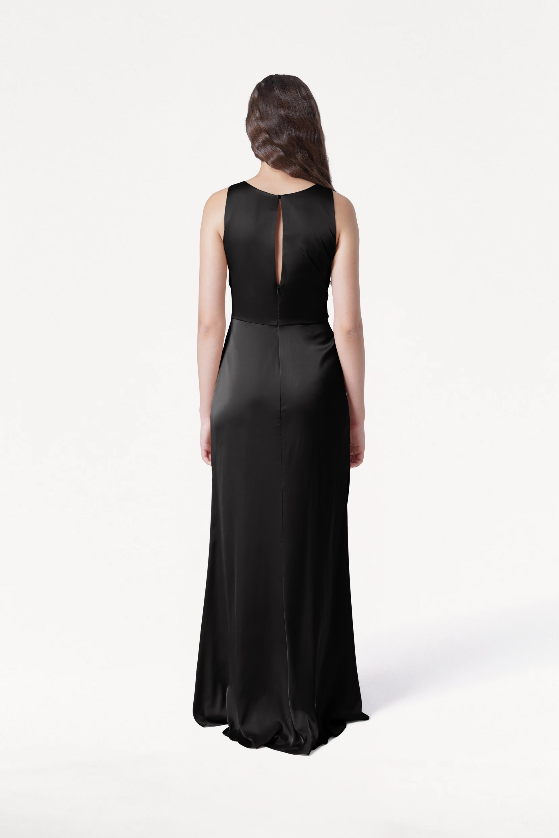 A woman with long brown hair models the TH&TH Nala Bridesmaid Dress in Black Satin, a sleeveless floor-length gown with Grecian-style pleating, standing with her back to the camera against a plain white background.