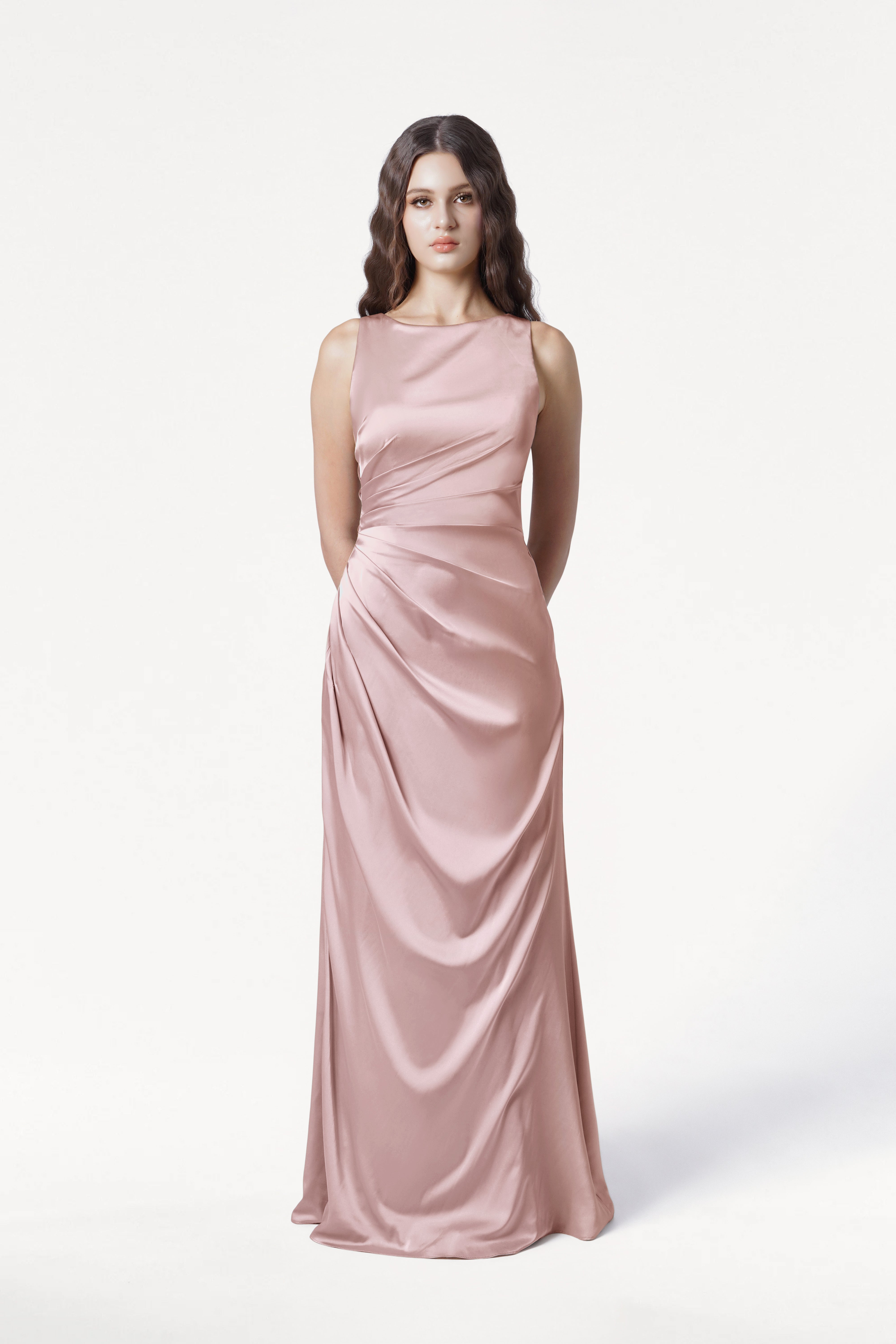 A woman with long, wavy dark hair wears the TH&TH Nala Bridesmaid Dress in Smoked Blush Satin—a sleeveless, floor-length gown with Grecian-style pleating and gentle draping—shown against a plain white background. Perfect for modern weddings.