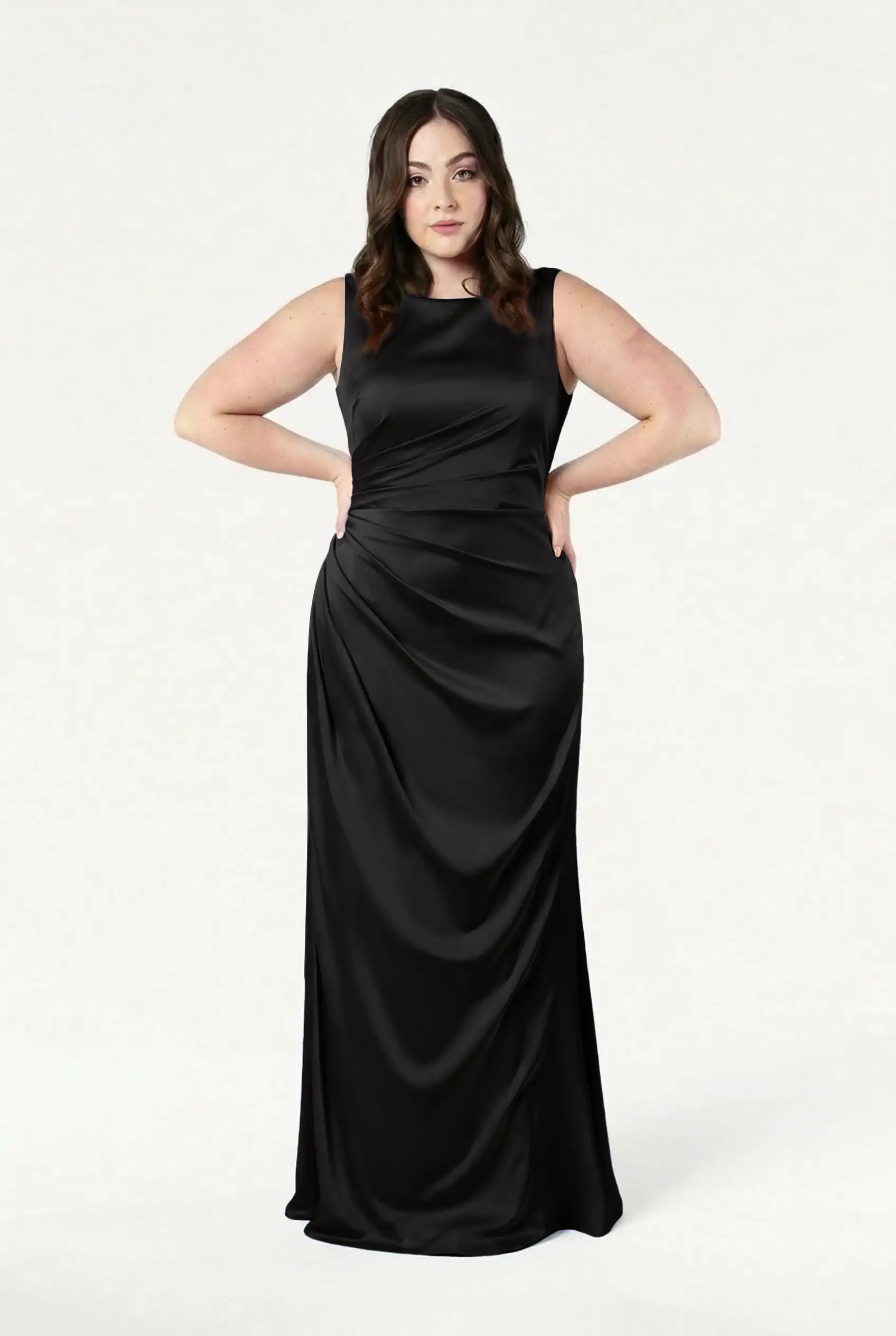 A woman with long brown hair stands confidently against a white background, wearing the TH&TH Nala Bridesmaid Dress in Black Satin—an inclusive, floor-length gown featuring ruched details and Grecian-inspired pleating.