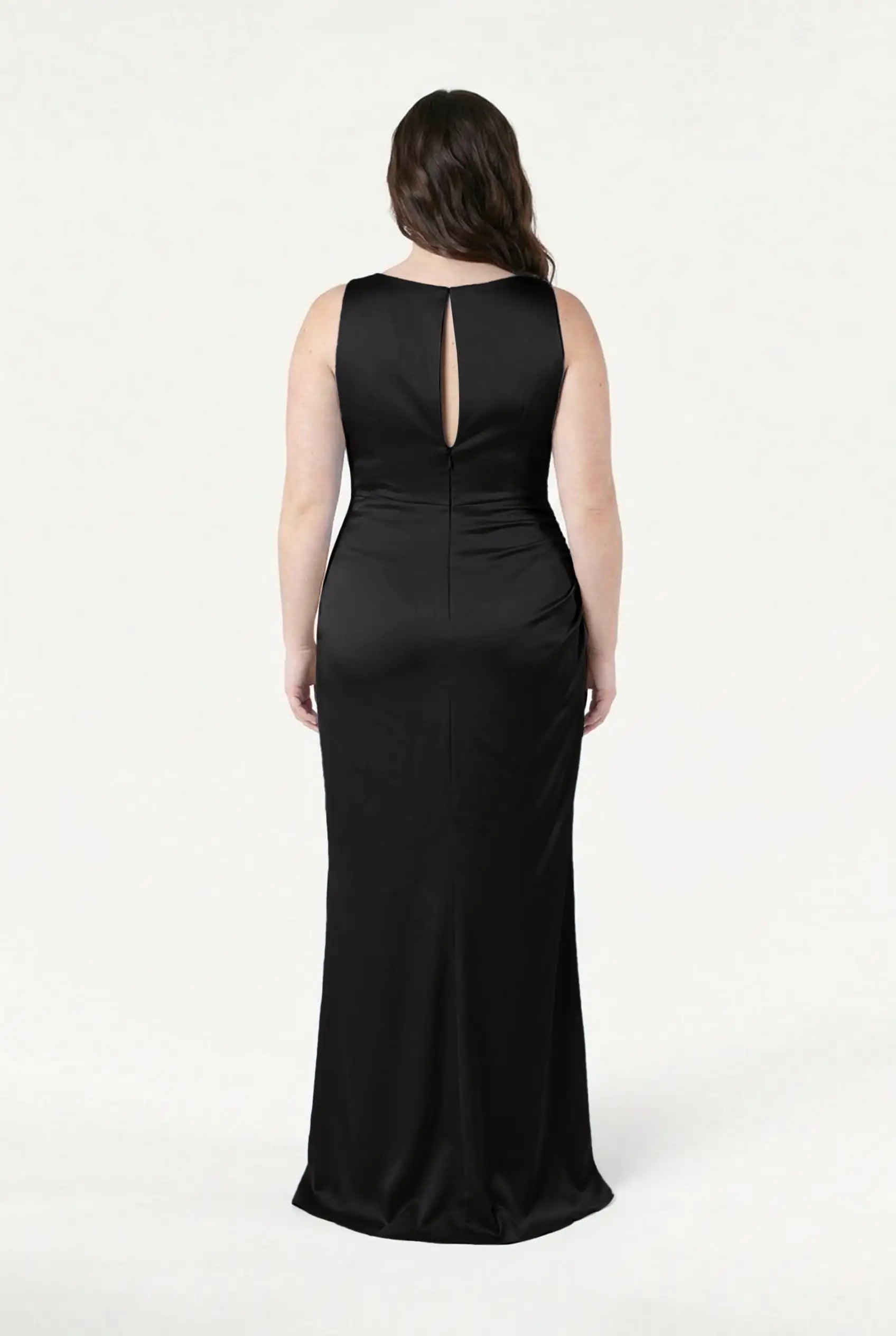 A woman with long dark hair models the TH&TH Nala Bridesmaid Dress in Black Satin, a sleeveless, floor-length gown with a keyhole back detail. She stands with her back to the camera against a plain white background.