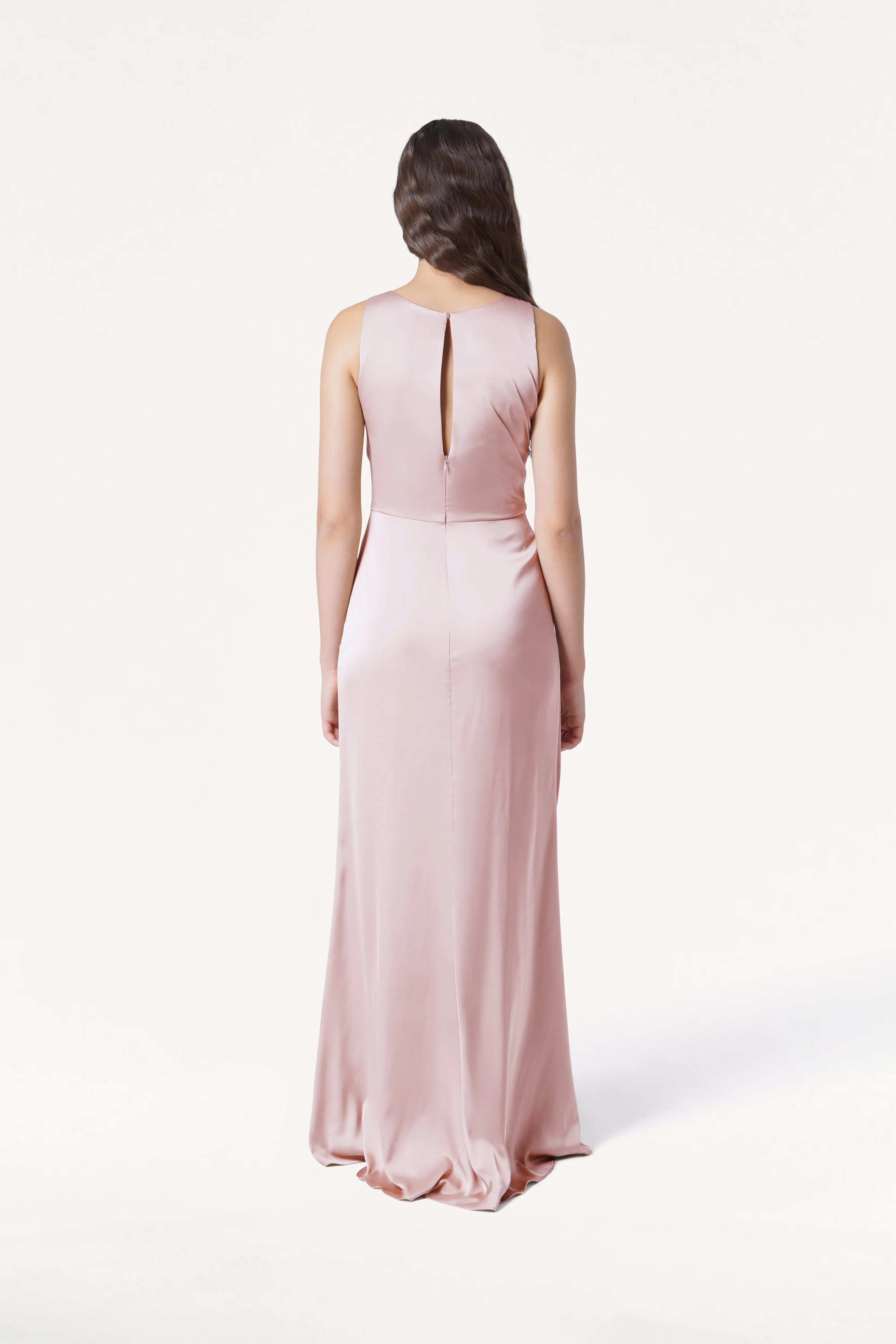 A woman in the TH&TH Nala Bridesmaid Dress in Blush Pink Satin stands facing away, showcasing Grecian-inspired pleating and a keyhole back. This modern gown is ideal for contemporary weddings, set against a simple white background.
