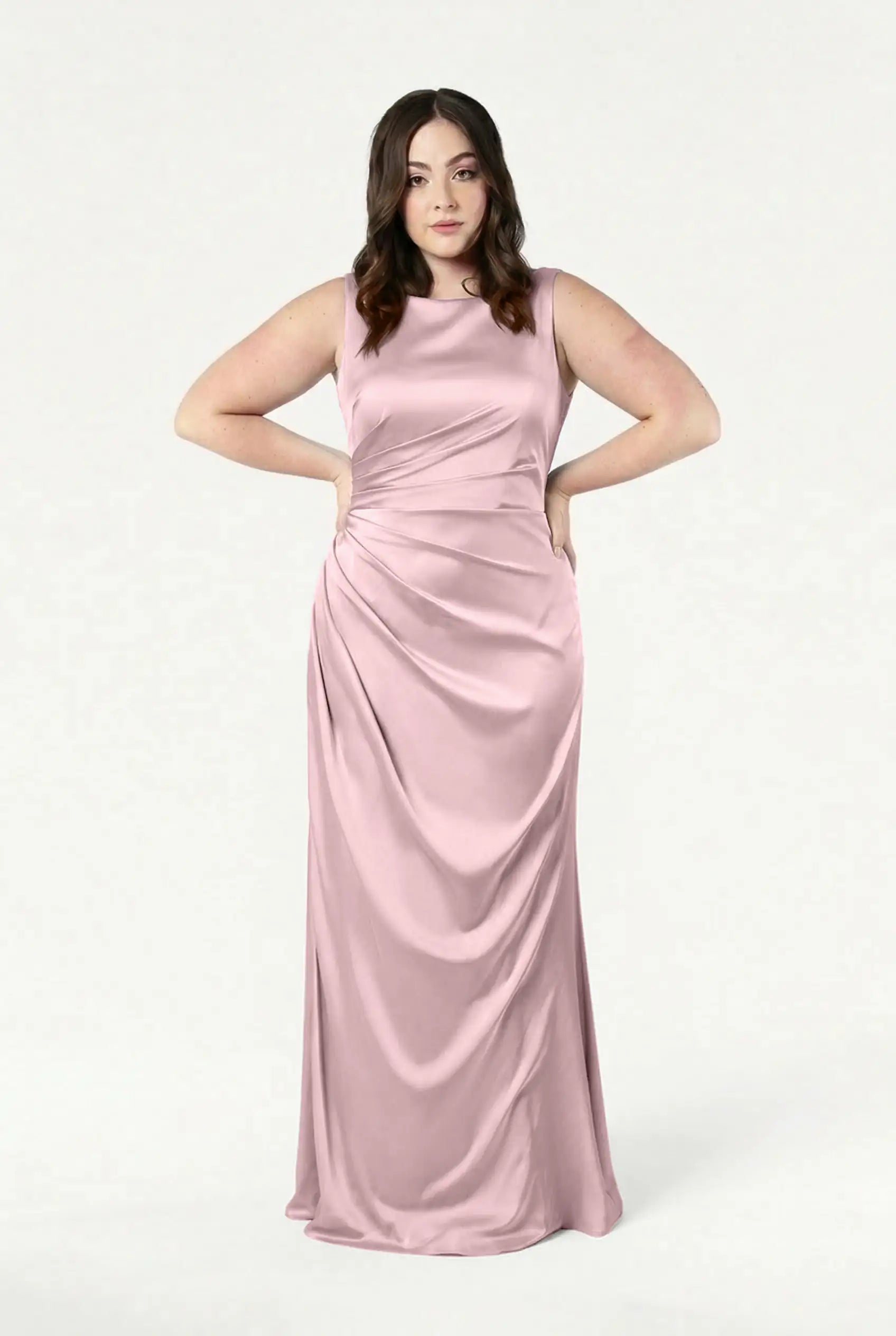 A woman with long dark hair stands with hands on hips, wearing the TH&TH Nala Bridesmaid Dress in Blush Pink Satin, a sleeveless, floor-length gown with Grecian pleating and ruching—ideal for modern weddings—against a plain white background.