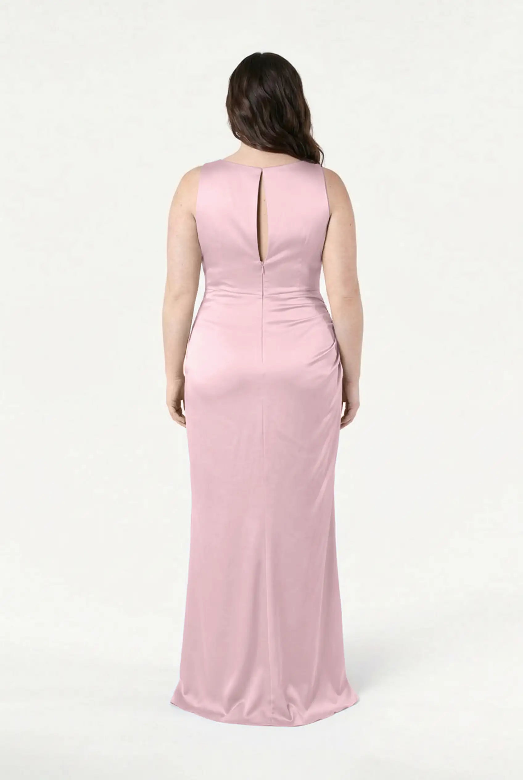 A woman faces away, wearing the TH&TH Nala Bridesmaid Dress in blush pink satin. This form-fitting, floor-length gown features Grecian-inspired pleating and a keyhole back, ideal for modern weddings. Background is plain and light.