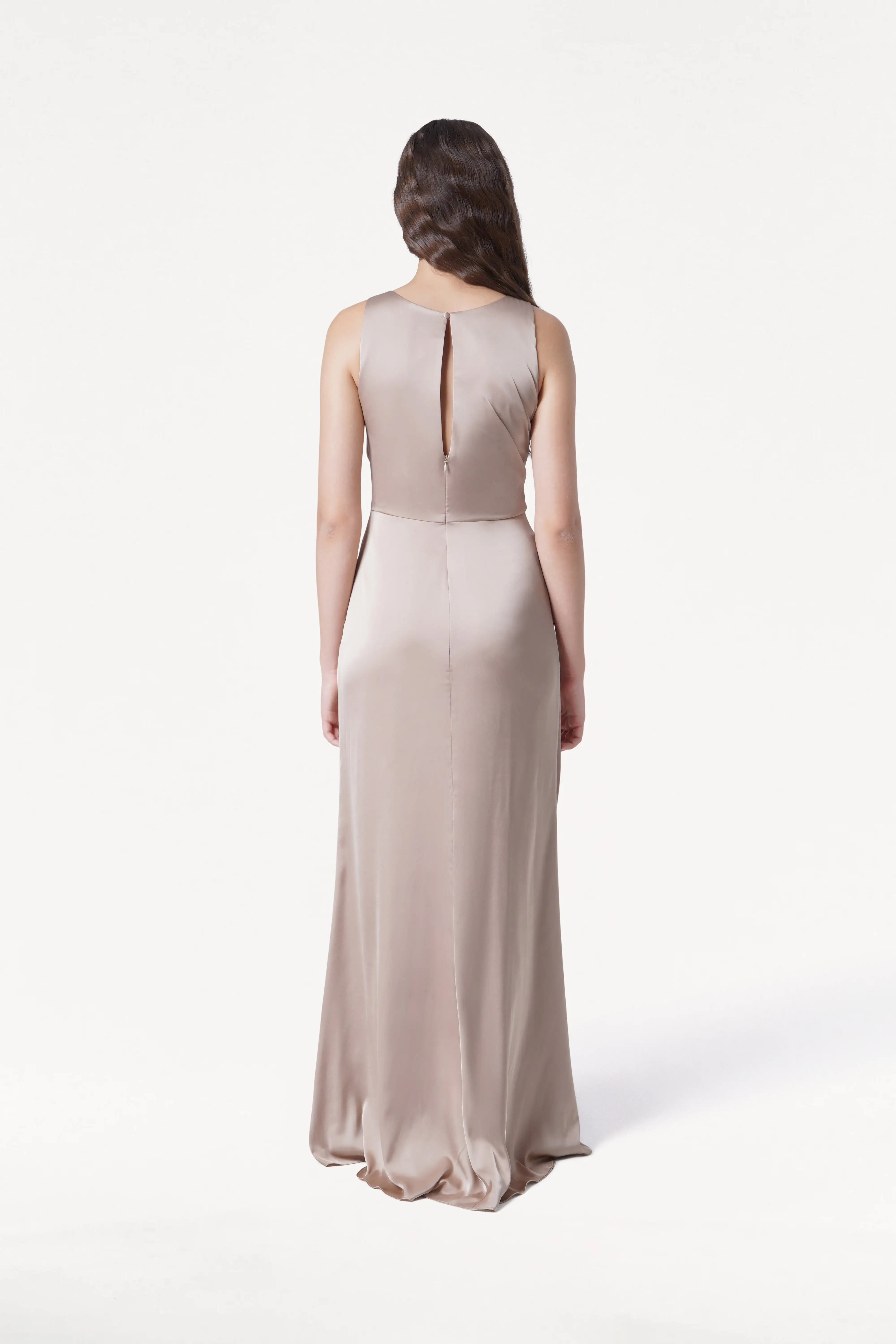 A woman with long brown hair poses against a white background, wearing the TH&TH Nala Bridesmaid Dress in Champagne Sky Satin—a sleeveless, floor-length gown with Grecian pleating and a subtle keyhole back detail.