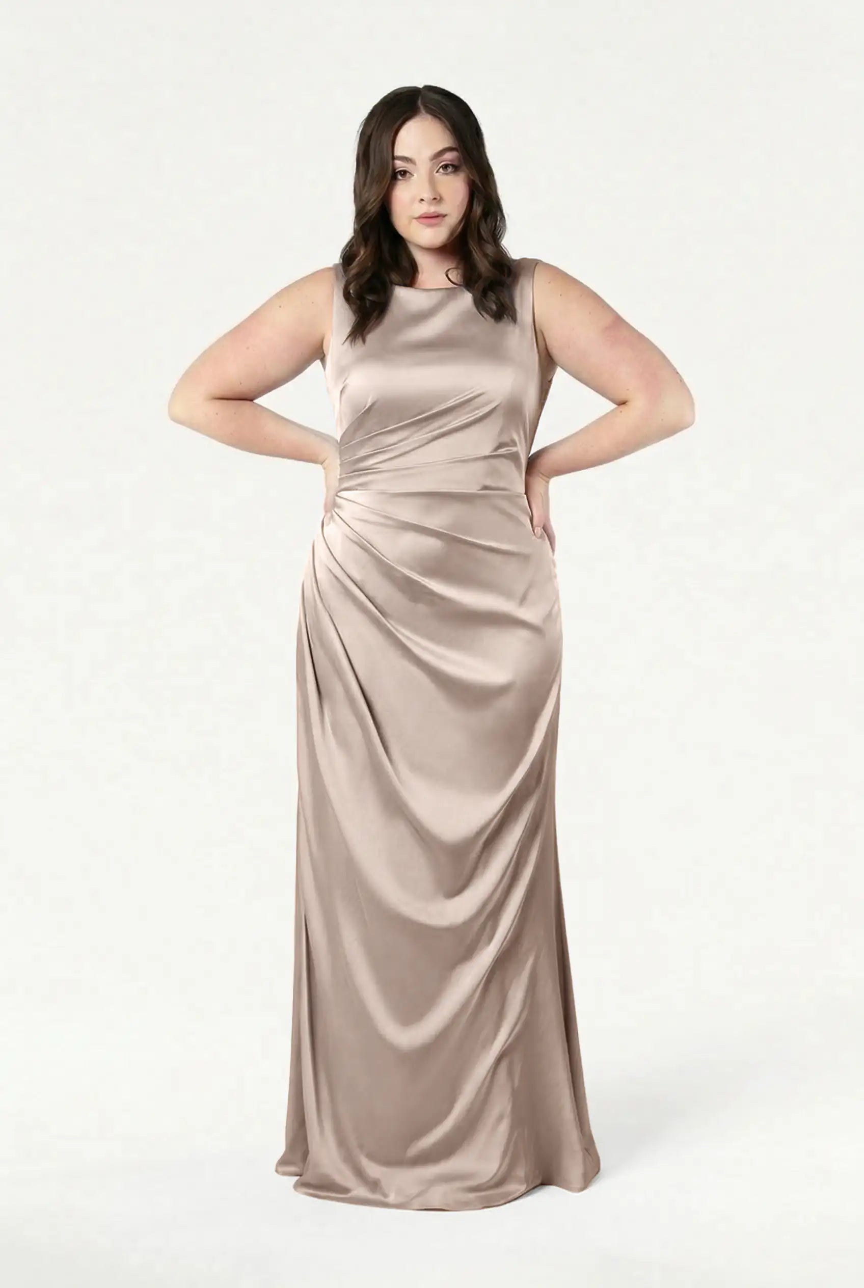 A woman with long, dark wavy hair poses against a white background wearing the TH&TH Nala Bridesmaid Dress in Champagne Sky Satin—a sleeveless, floor-length gown featuring Grecian-inspired pleating at the waist.