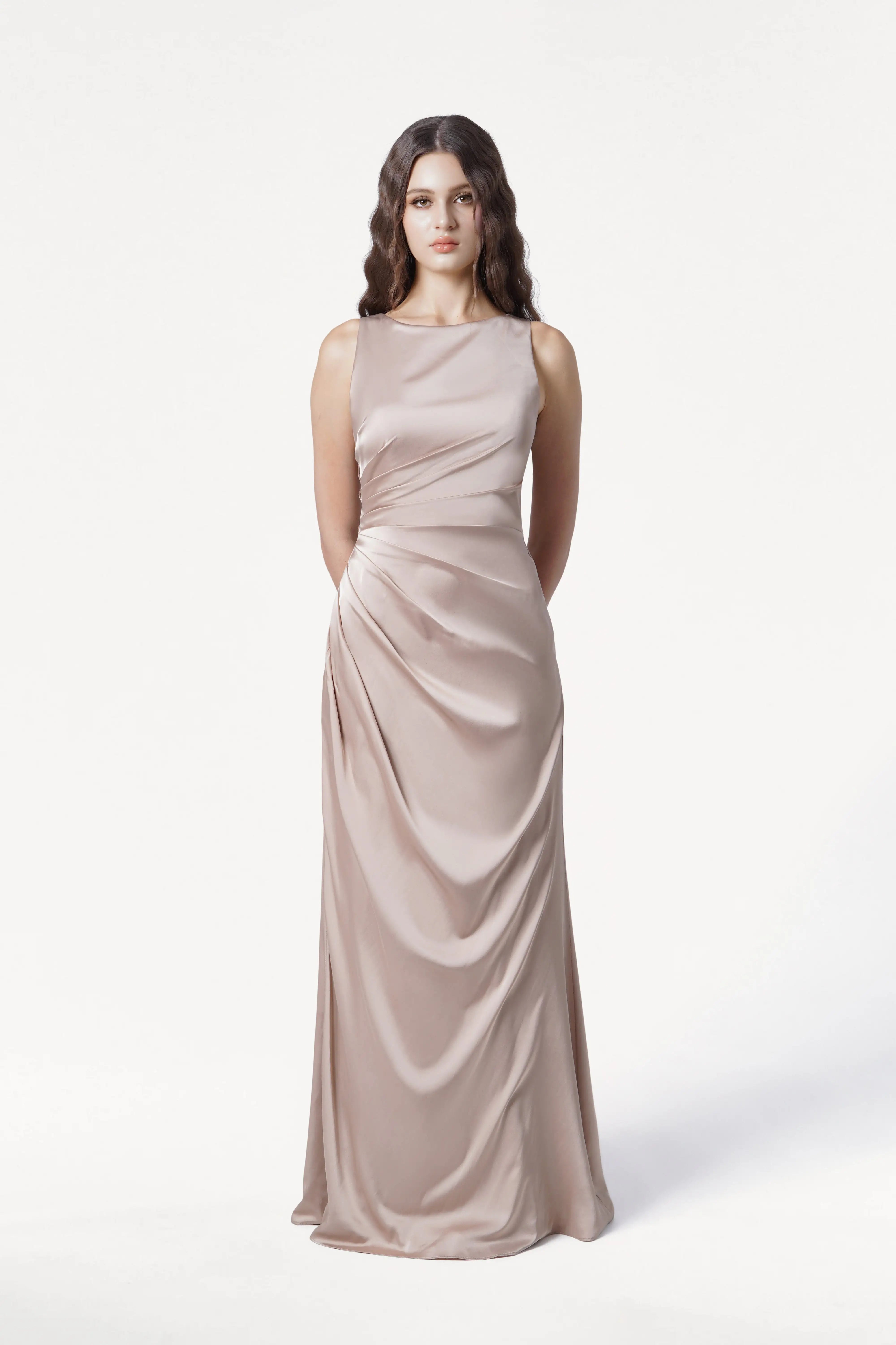 A woman with long wavy hair stands against a white background, wearing the TH&TH Nala Bridesmaid Dress in Champagne Sky Satin. The sleeveless, floor-length gown features Grecian-inspired pleating. Her arms are behind her back and she has a neutral expression.