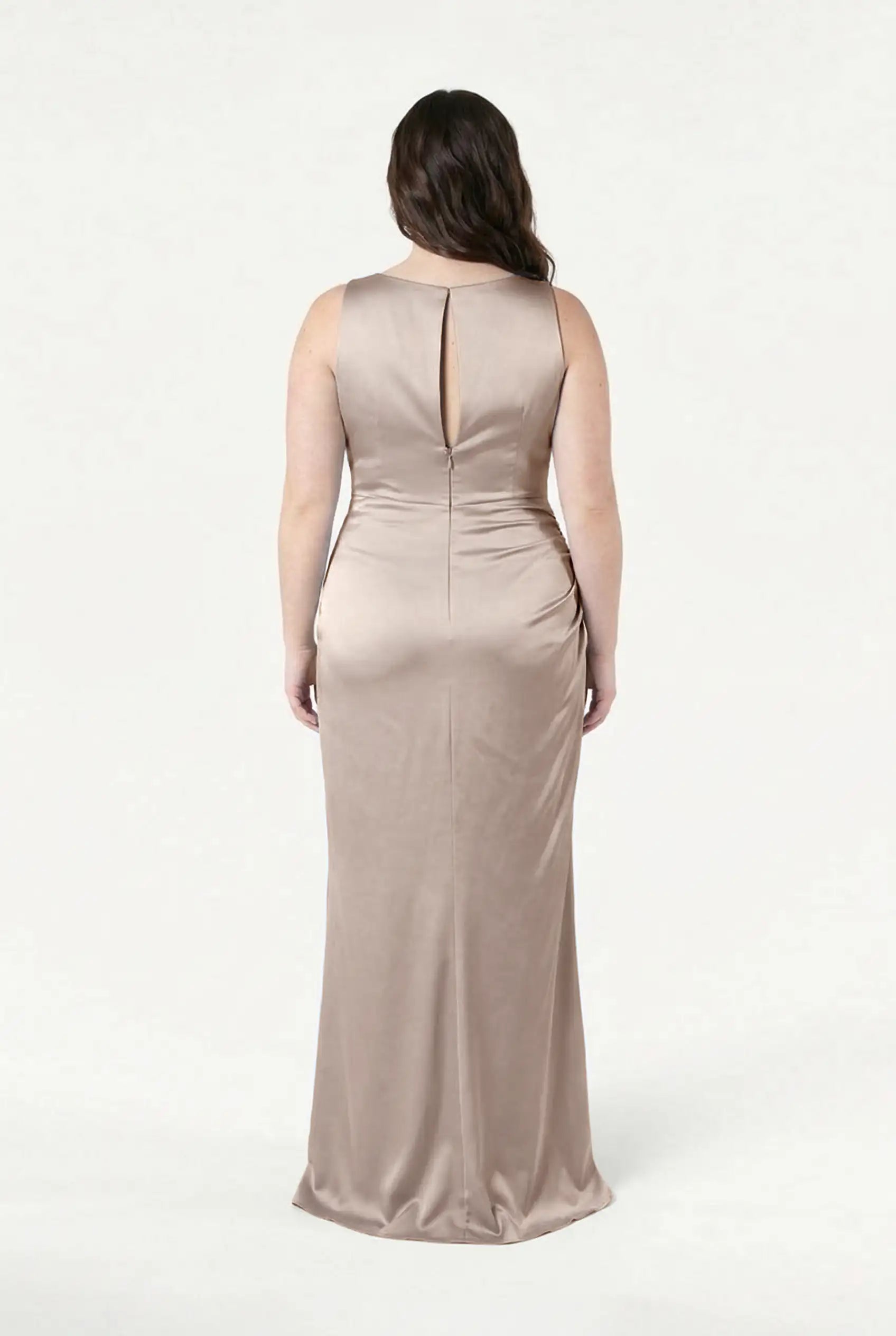 A person stands facing away, wearing the Nala Bridesmaid Dress in Champagne Sky Satin by TH&TH—a sleeveless, floor-length gown with Grecian-inspired pleats and a small keyhole back—set against a plain white background.