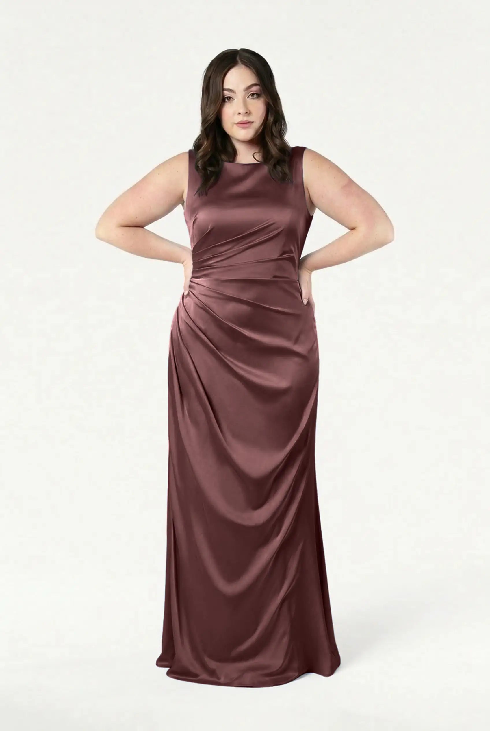 A woman stands confidently with her hands on her hips, wearing the TH&TH Nala Bridesmaid Dress in chocolate brown satin, a sleeveless, floor-length gown with gentle Grecian-inspired pleating on one side, against a plain white background.