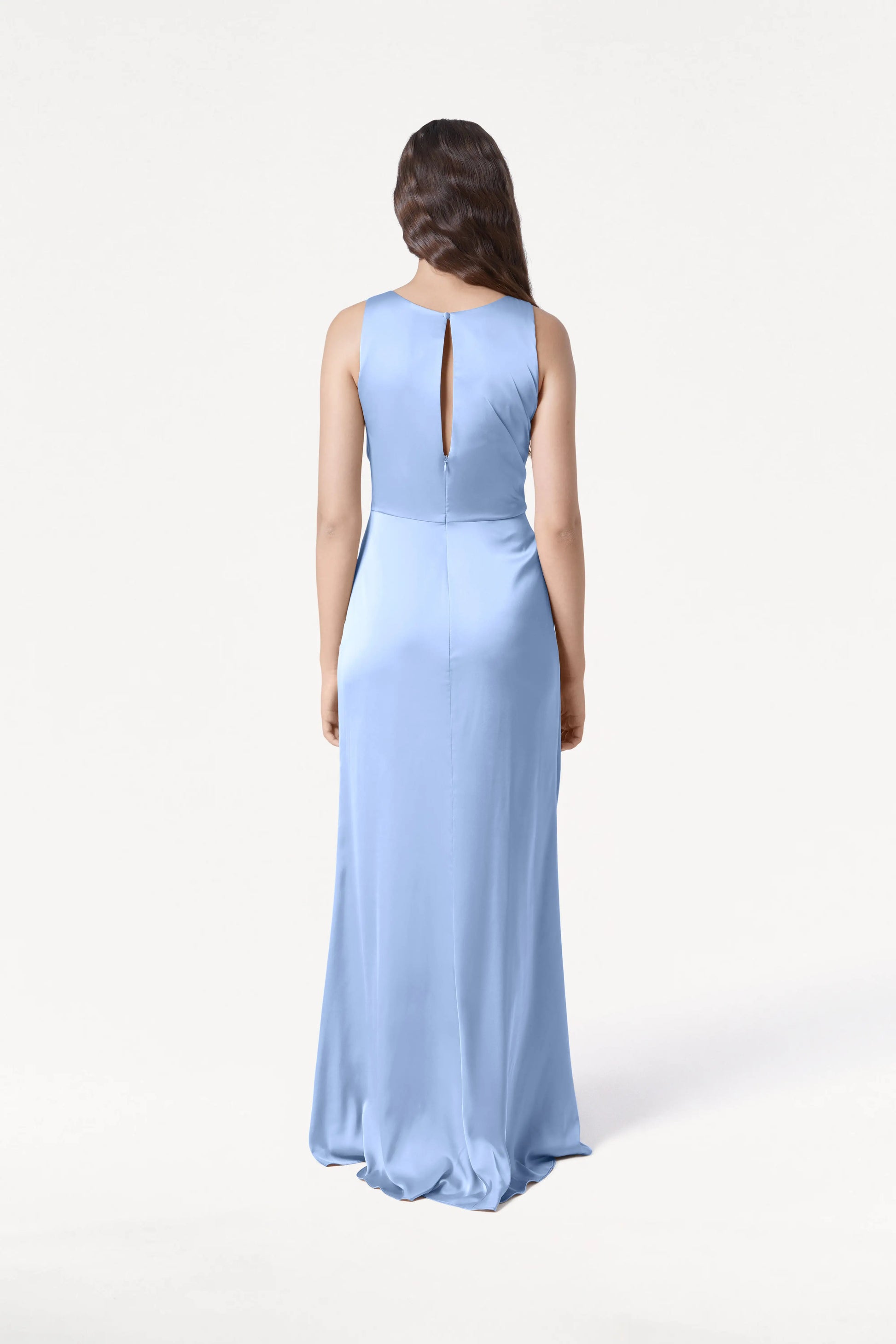 A woman with long brown hair faces away, wearing the TH&TH Nala Bridesmaid Dress in dusty blue satin—a size-inclusive, floor-length gown with Grecian-inspired pleating and a keyhole back—against a plain white background.