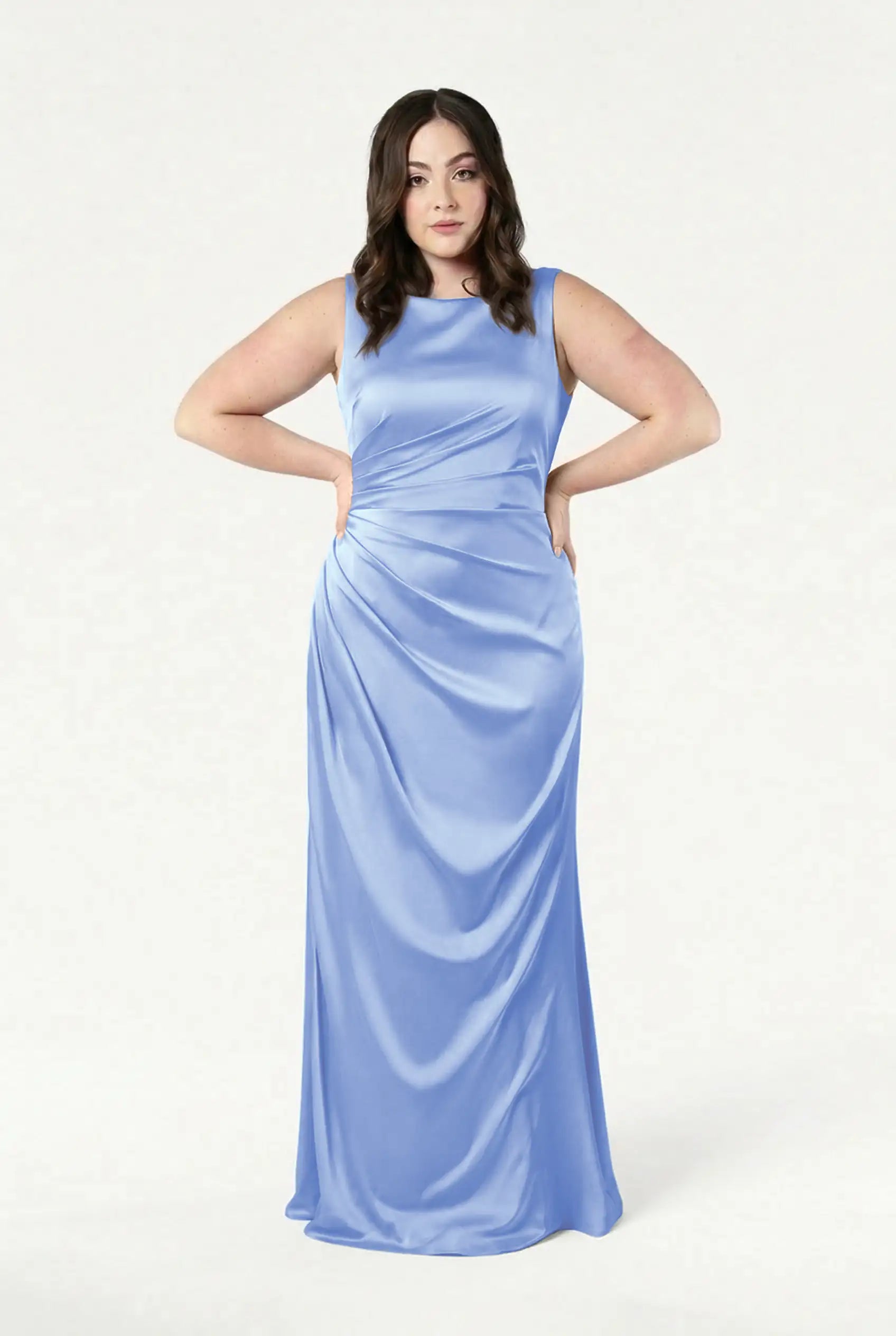 A woman poses with her hands on her hips in the TH&TH Nala Bridesmaid Dress, a sleeveless, floor-length gown in dusty blue satin, featuring Grecian-inspired pleating, against a plain white background.